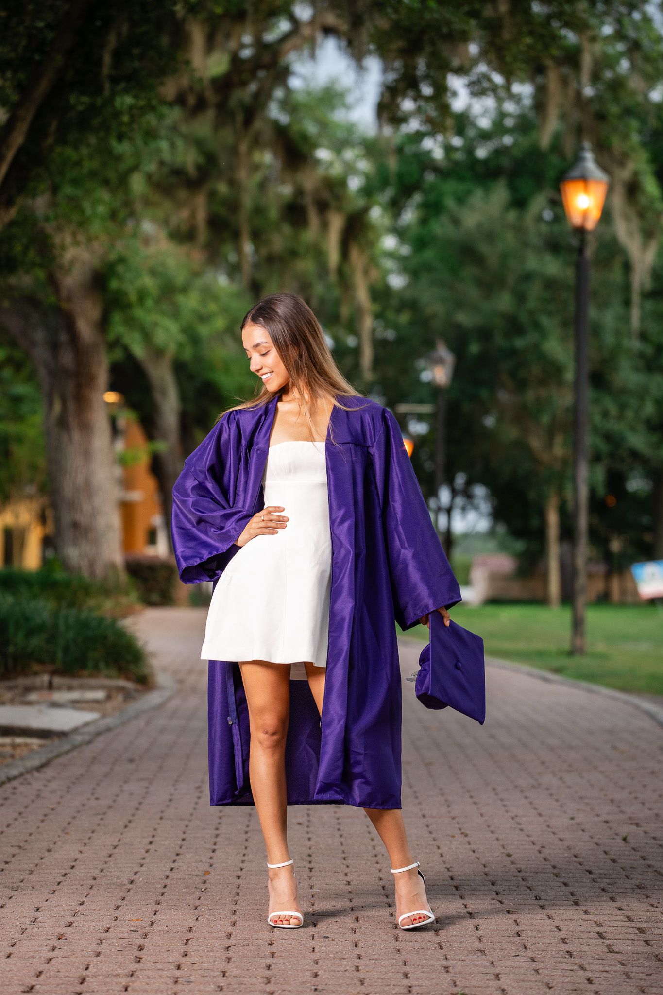 Graduate in purple cap and gown walking on brick path under moss-covered trees at dusk.