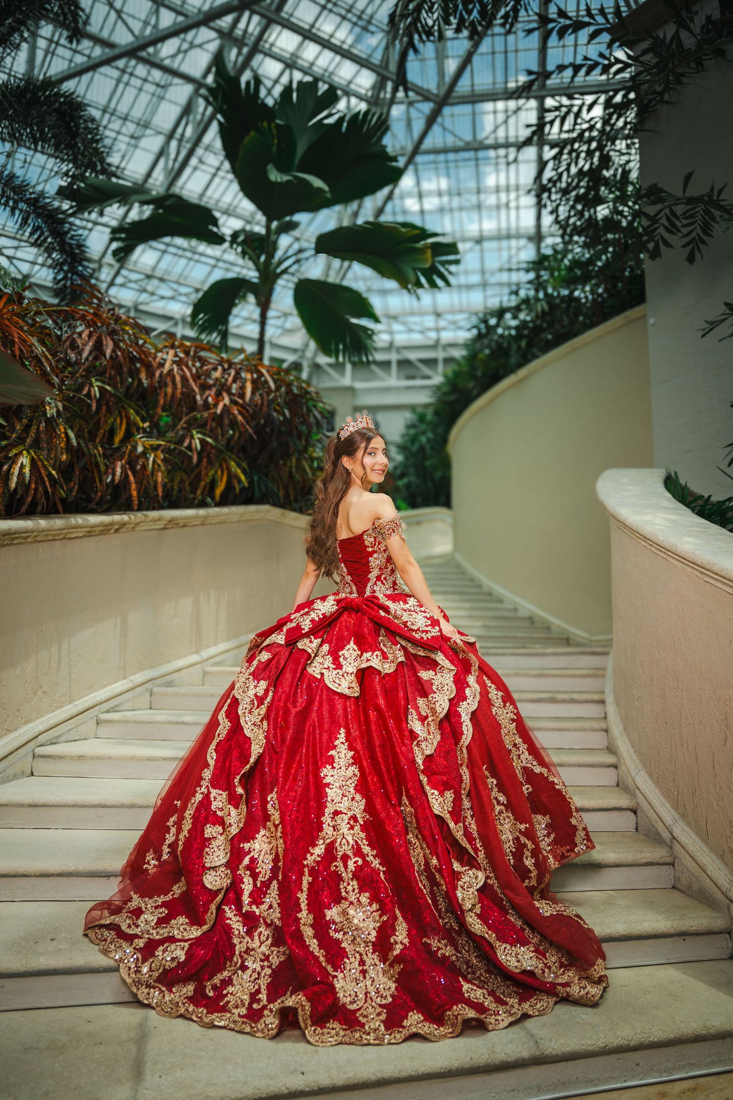 Young woman in red and gold ball gown on stairs in a lush greenhouse, perfect for quinceañera inspiration.