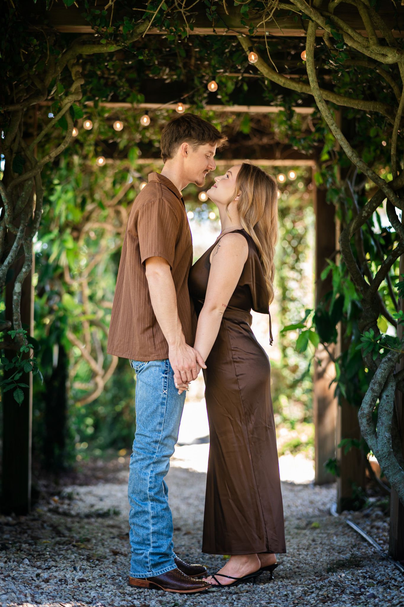 A couple holds hands and gazes at each other under a vine-covered pergola, dressed in coordinating brown outfits.