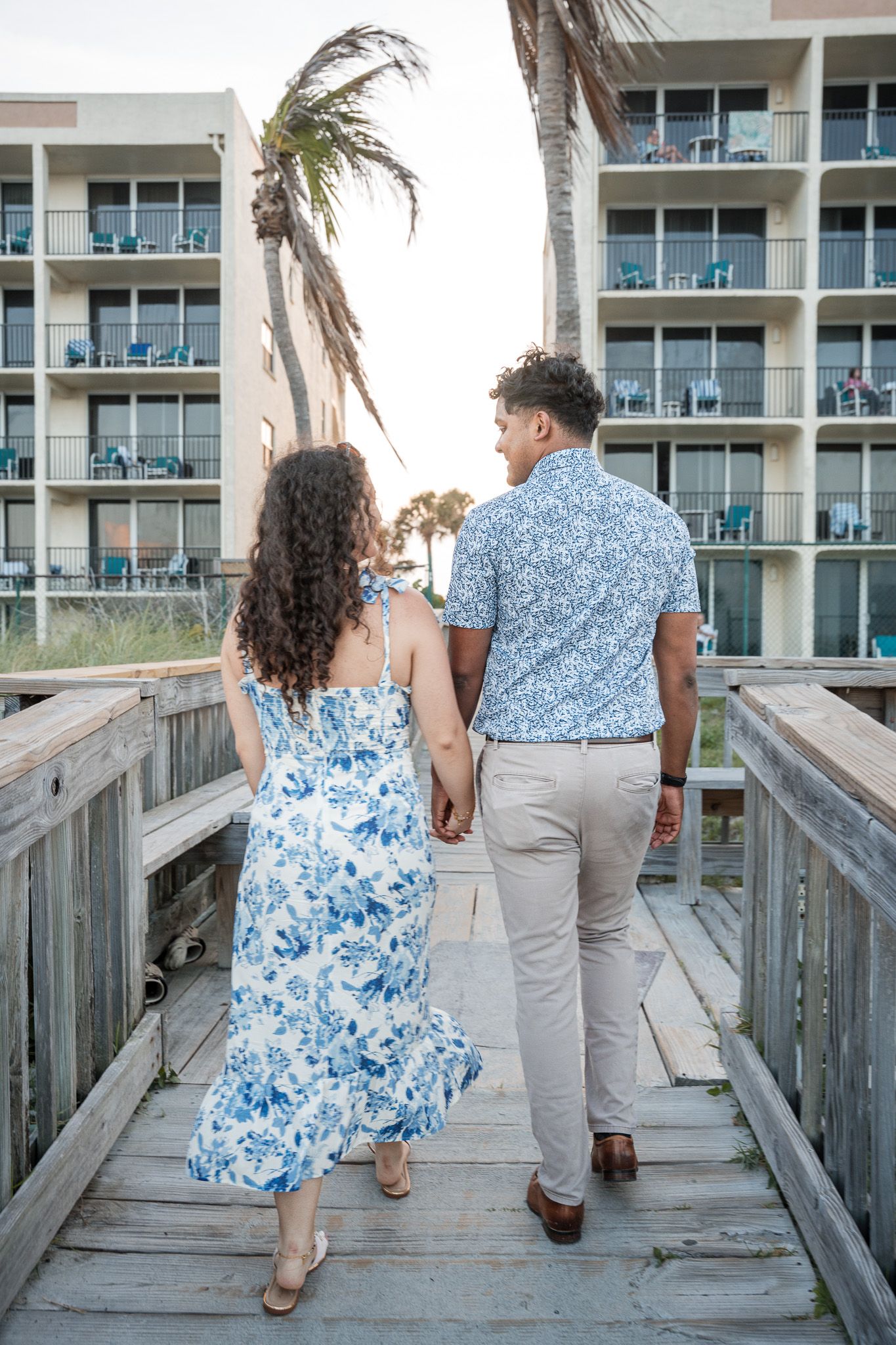 Couple holding hands walking away on wooden boardwalk toward beachfront hotel with palm trees at sunset.
