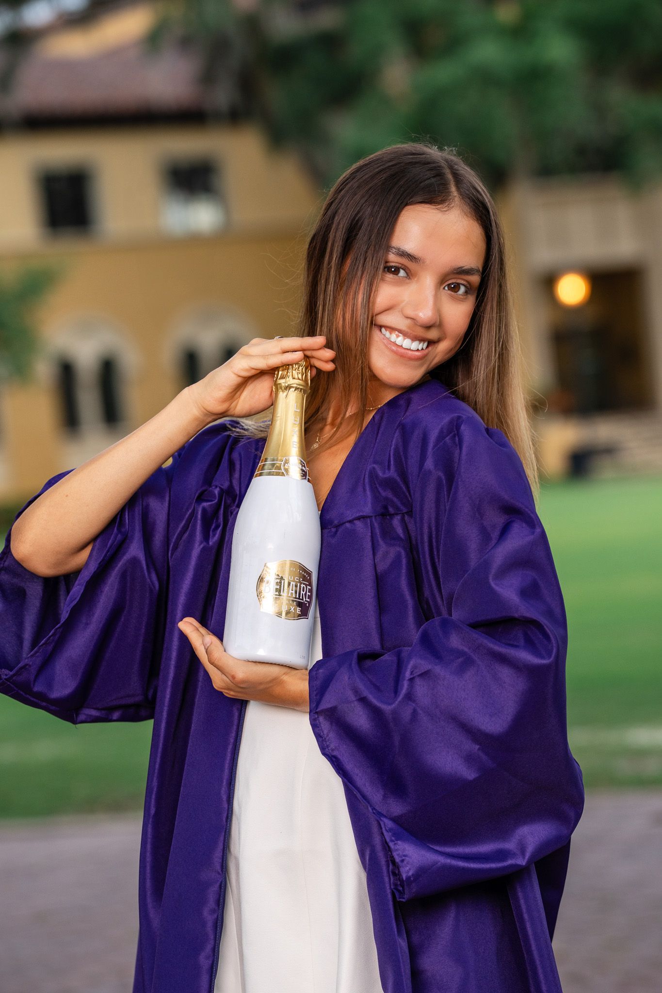 Happy female graduate in purple gown holds champagne bottle outdoors on college campus grounds.