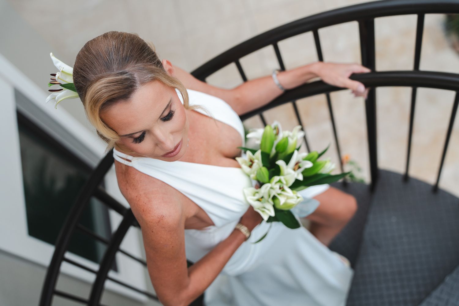 Elegant bride in white halter gown carries white lily bouquet while walking down spiral staircase.