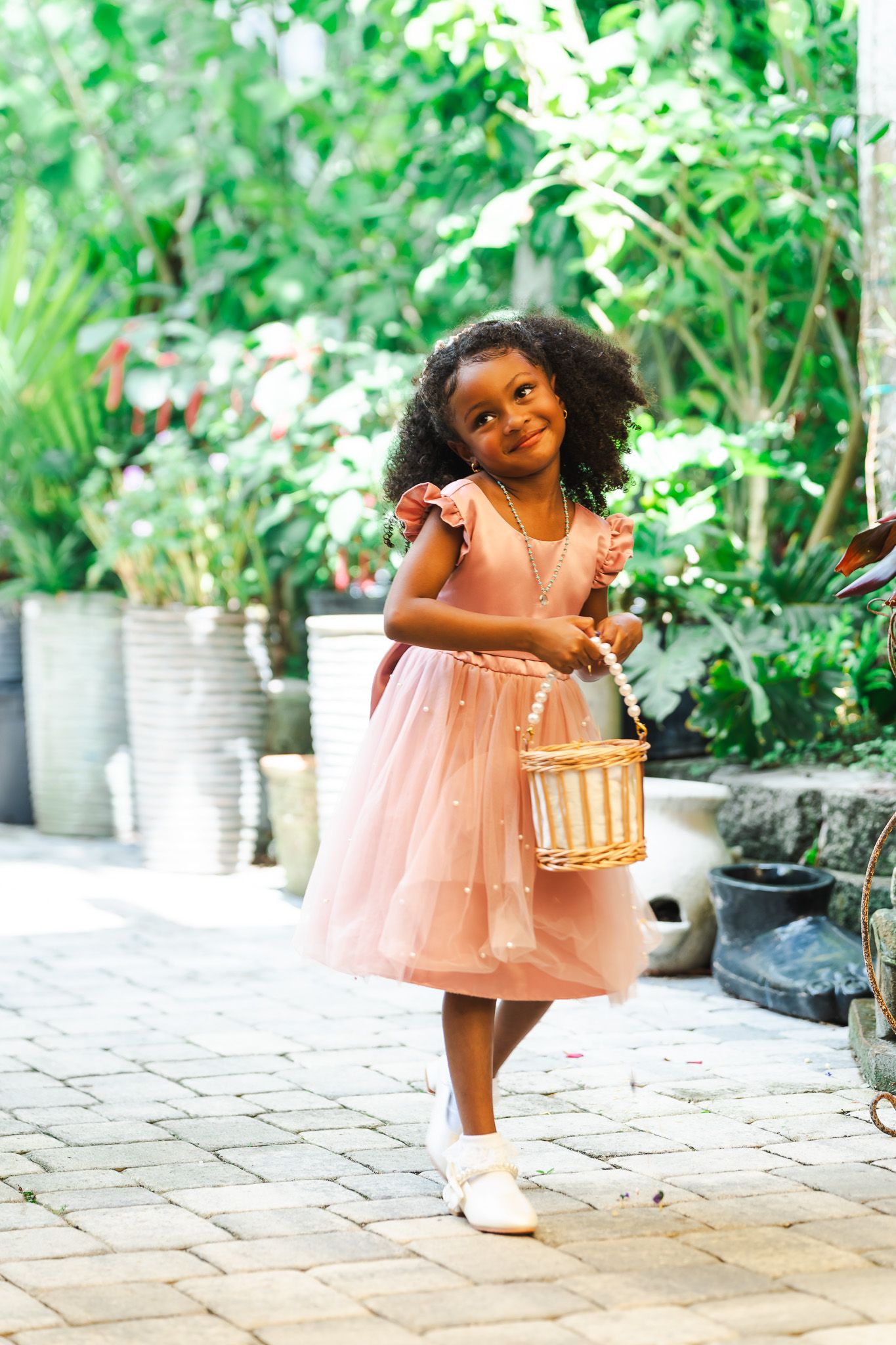 Adorable young flower girl in a pink tulle dress holding a wicker basket in a garden setting.
