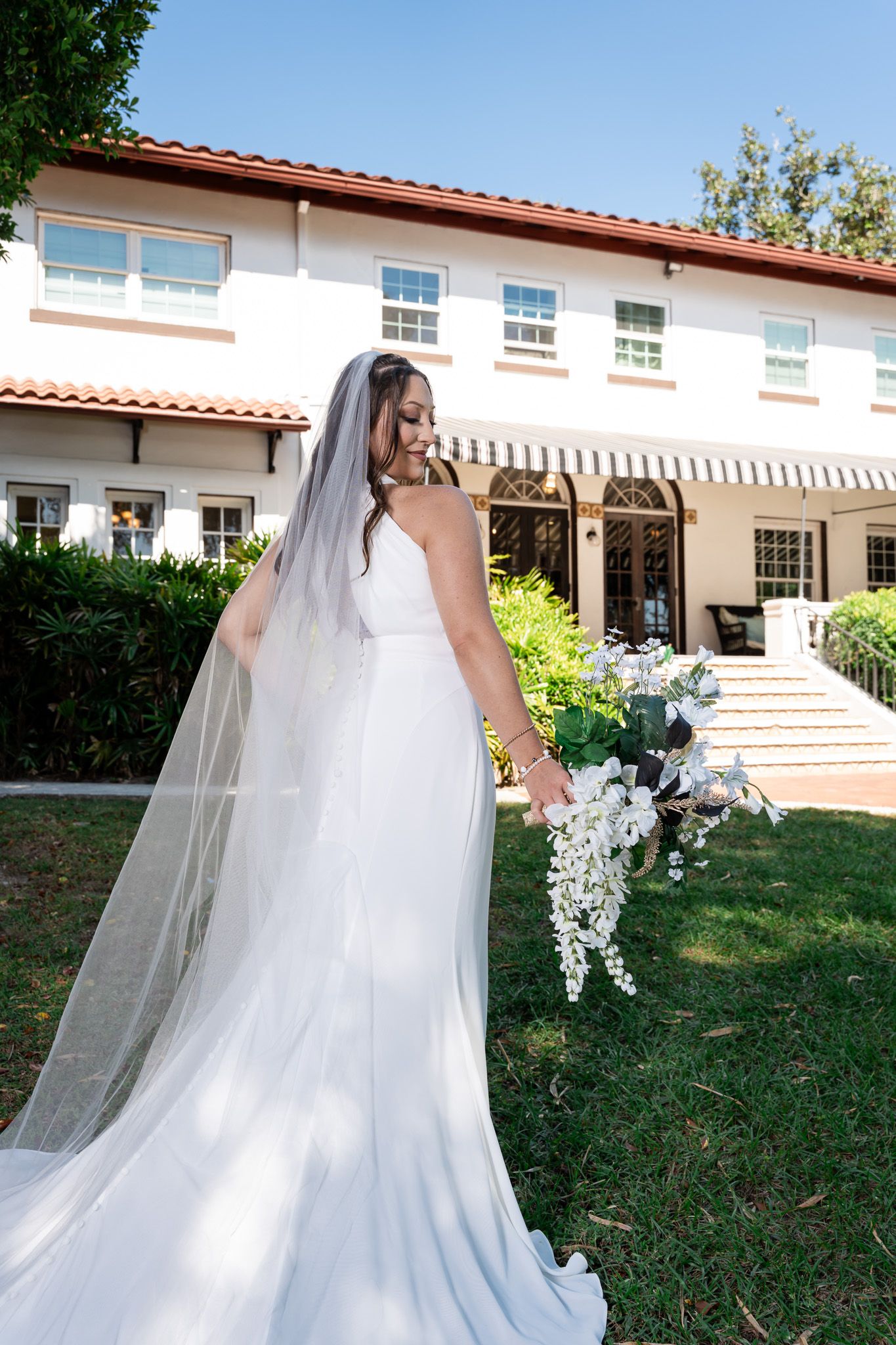 Bride in elegant white halter gown with cathedral veil holds cascading white bouquet outside venue.