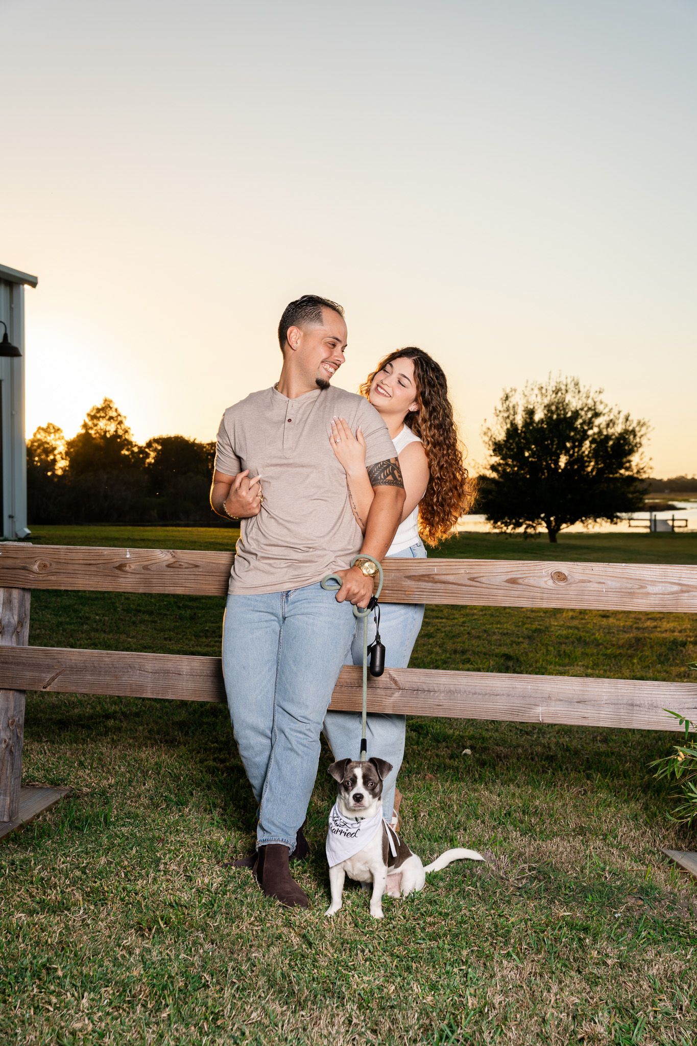Couple posing with their small dog on a leash by a wooden fence during a beautiful sunset session.