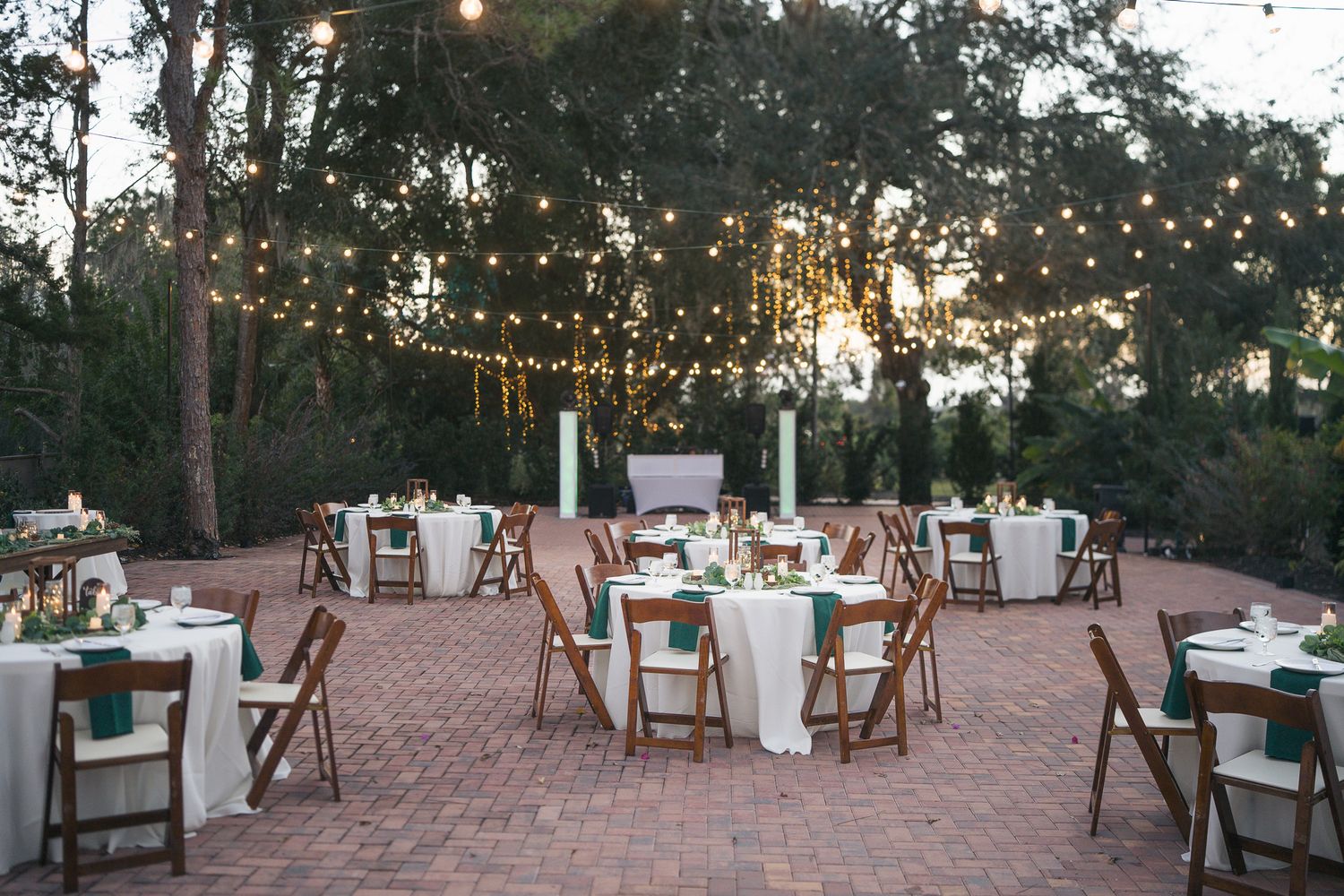 Outdoor wedding reception setup with round tables, wooden chairs, and string lights at sunset.