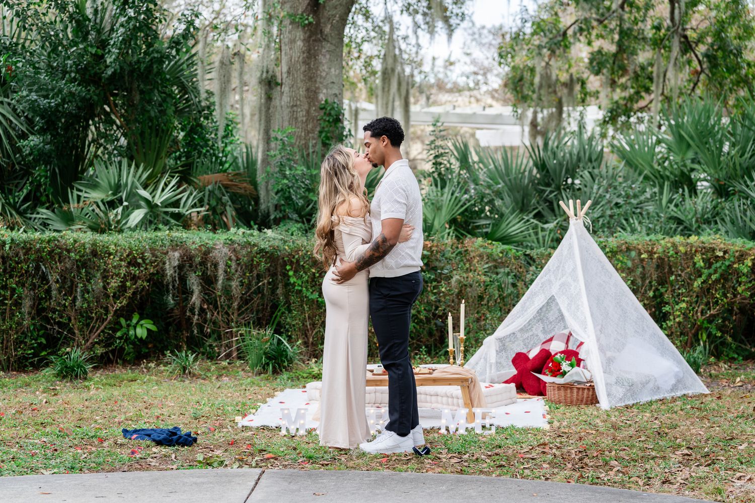 Newly engaged couple embracing near white teepee tent and picnic setup in tropical garden.
