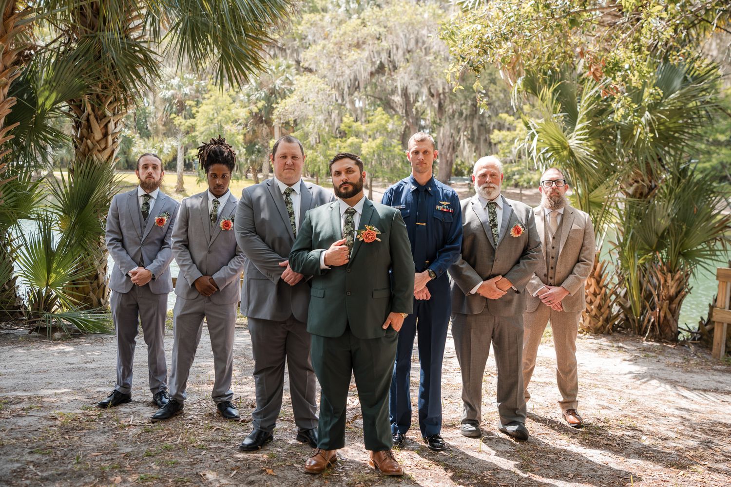 Groom in green suit poses with groomsmen in tan suits outdoors among palm trees.