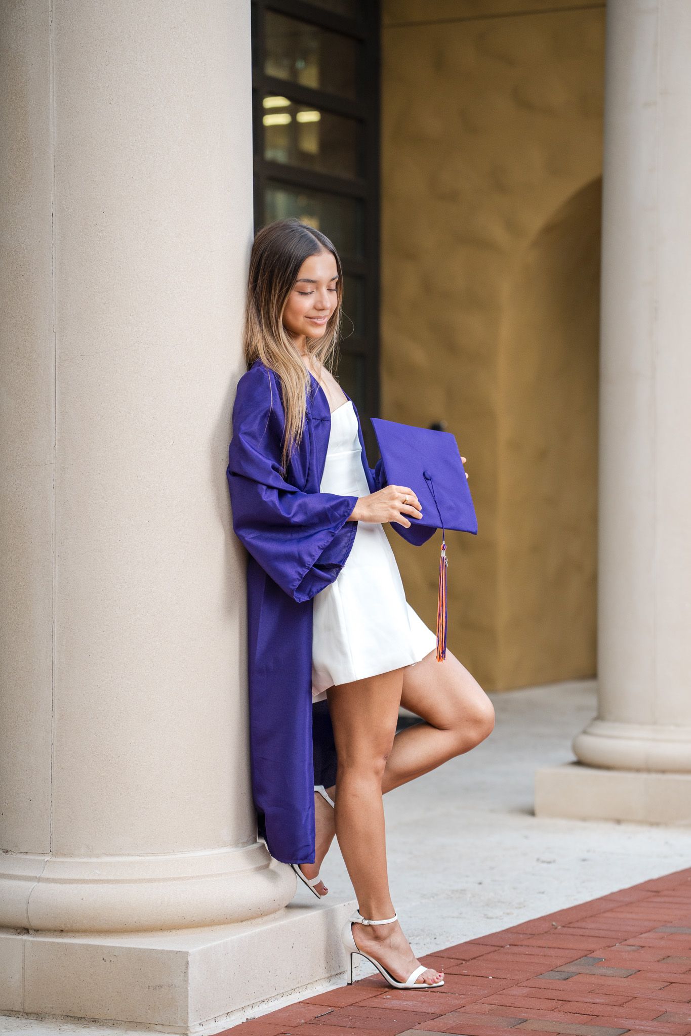 Young woman in purple graduation gown and cap leaning against column at university for senior photos.