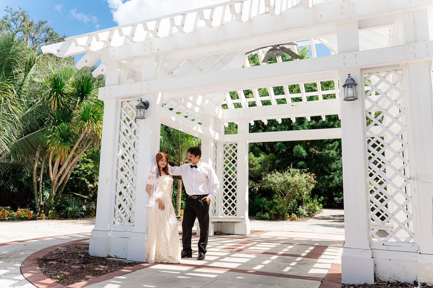 Bride and groom pose together under elegant white pergola archway in lush garden wedding venue.