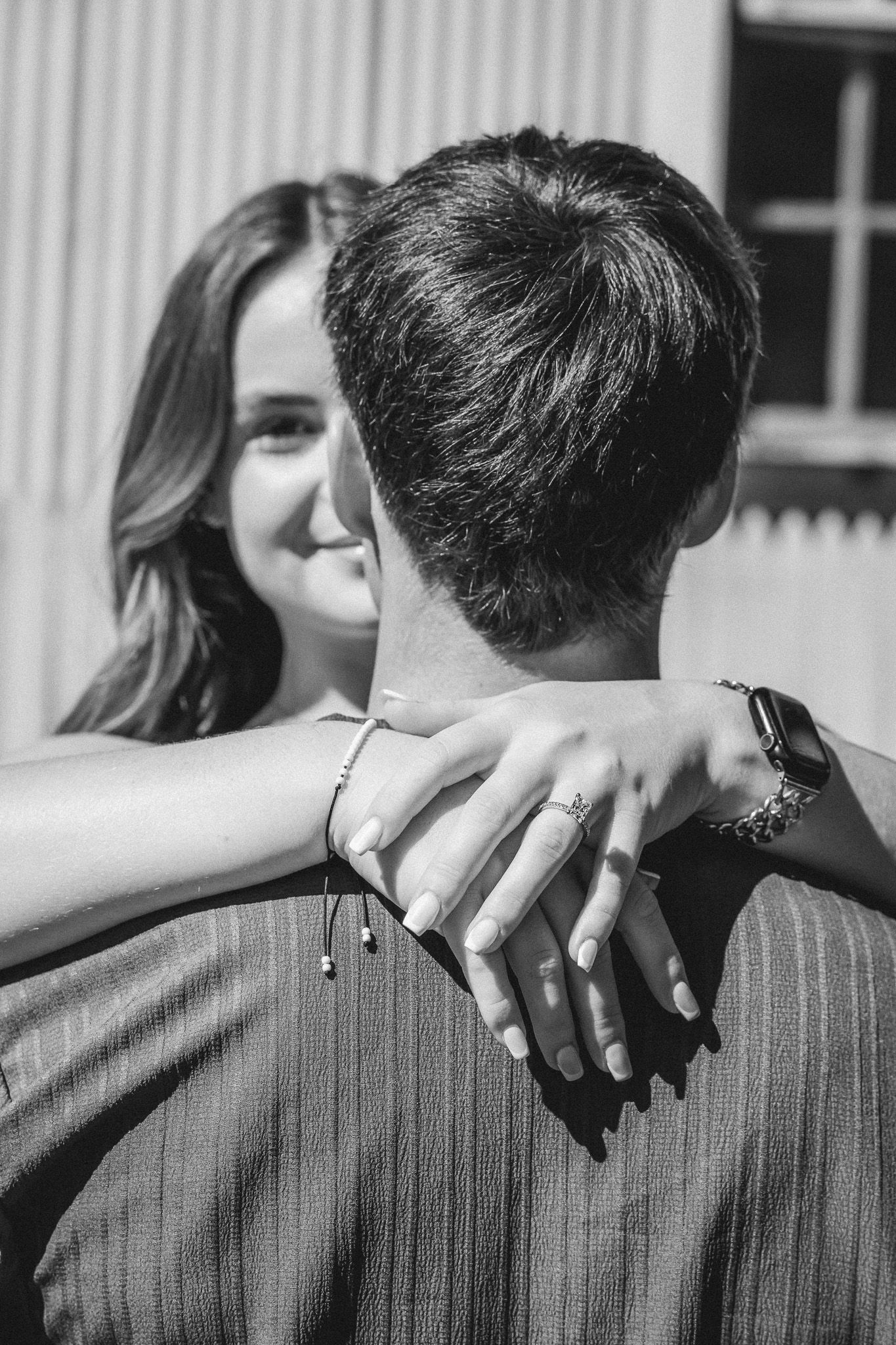 Black and white photo of a smiling woman embracing a man from behind, wearing a ring and bracelet.