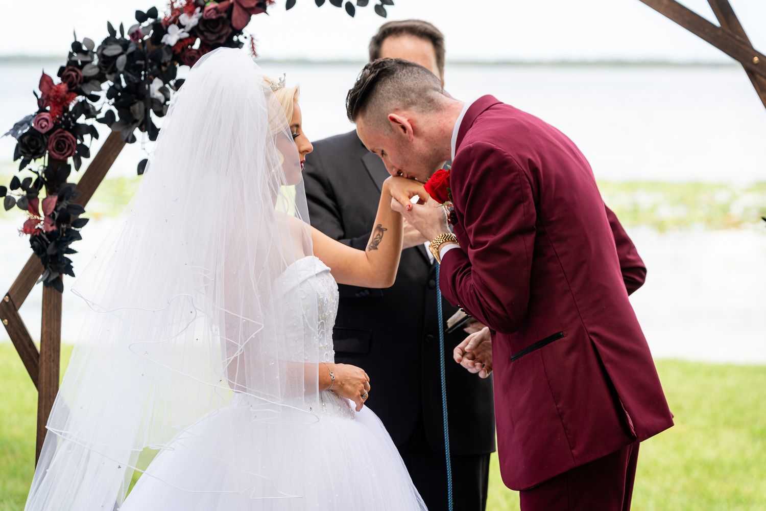 Groom in burgundy suit kisses bride's hand during outdoor wedding ceremony under floral arch by water.