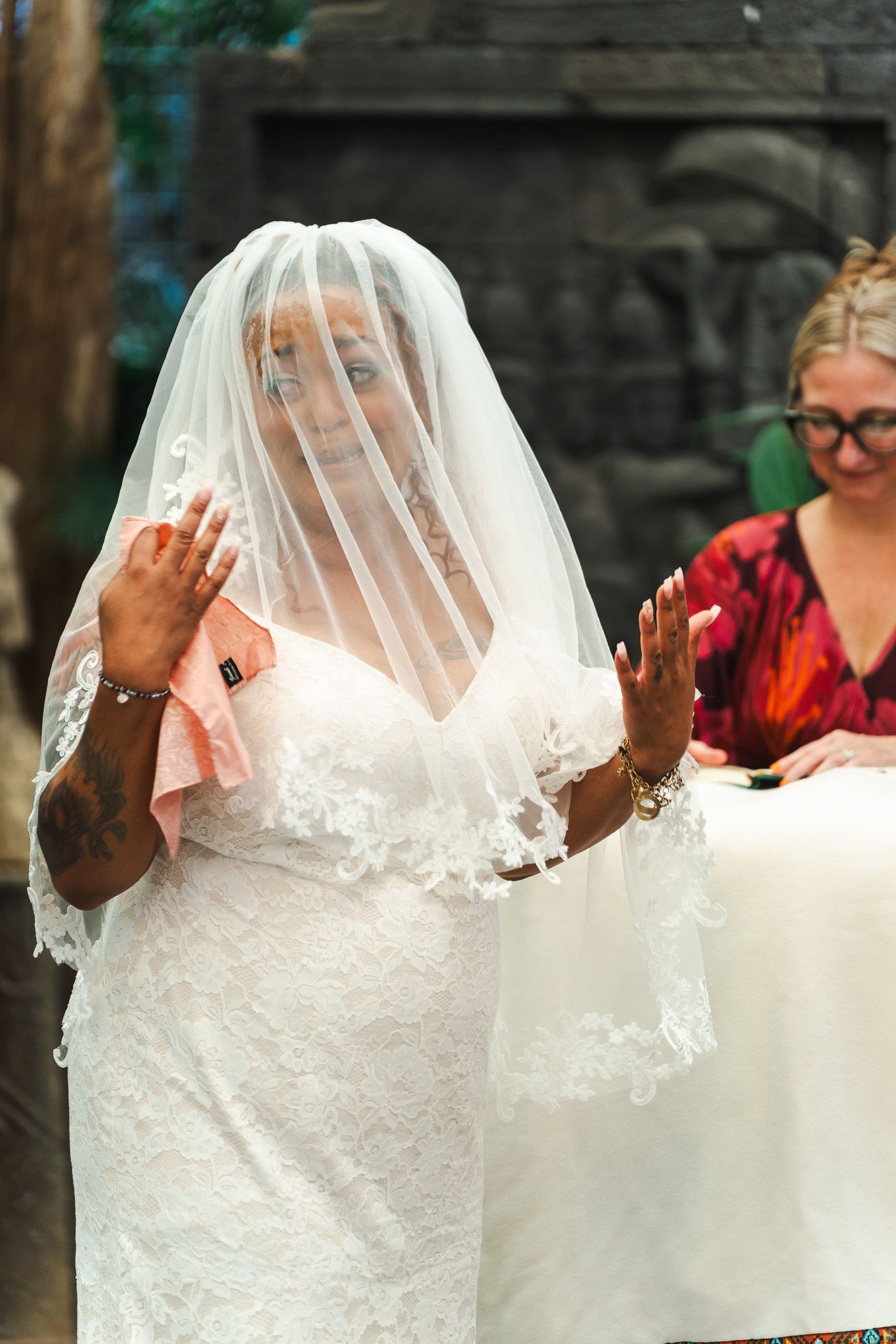 Emotional bride in lace gown and veil raises hands during outdoor wedding ceremony.