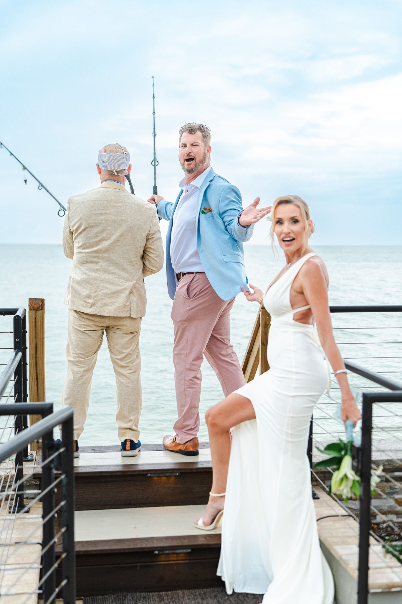 Groom gestures expressively on dock while older man fishes and bride laughs at seaside wedding.