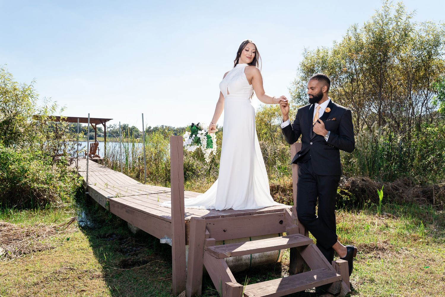 Bride and groom pose on a wooden dock by a lake, holding hands as bride stands elevated on steps.