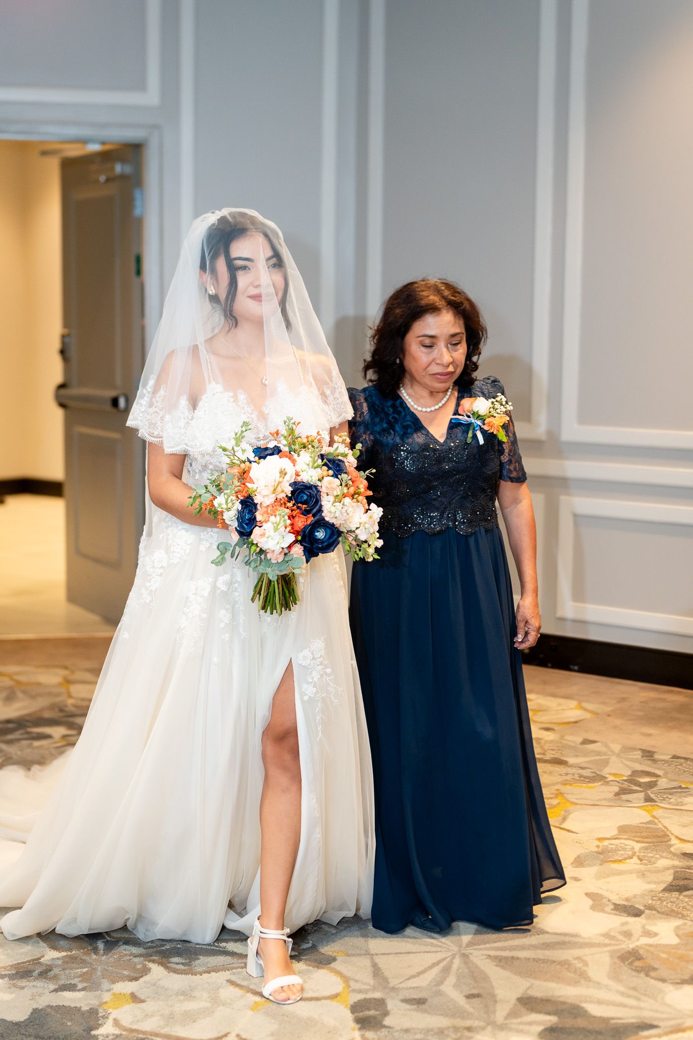 Bride in lace gown with veil walks with mother in navy dress during wedding ceremony procession.