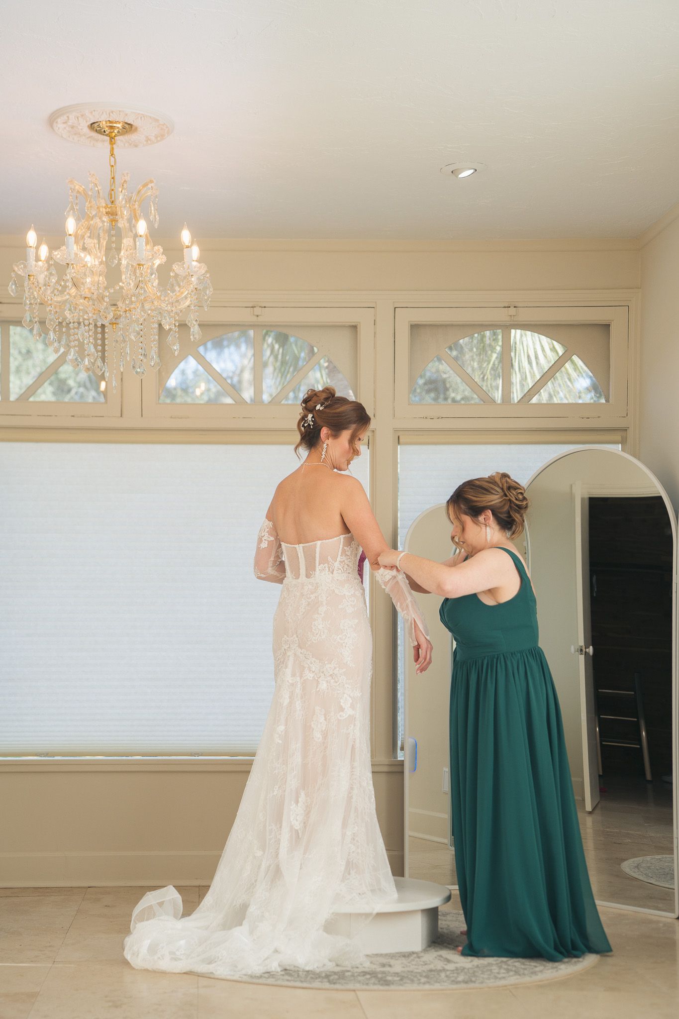 Bridesmaid in green dress helps bride button her lace wedding gown under chandelier.