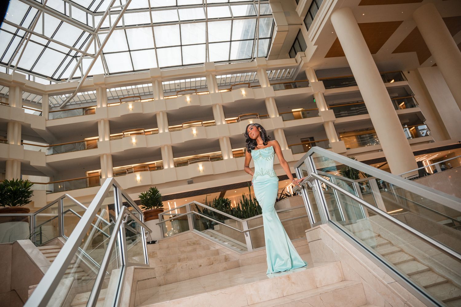Woman in mint green mermaid gown poses on stairs inside elegant atrium building with glass ceiling.