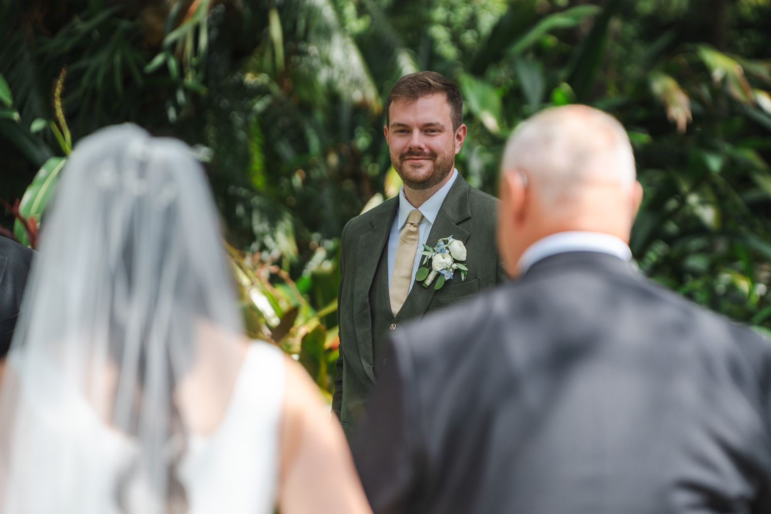 Groom smiles joyfully as bride approaches the altar at a lush tropical outdoor wedding ceremony.