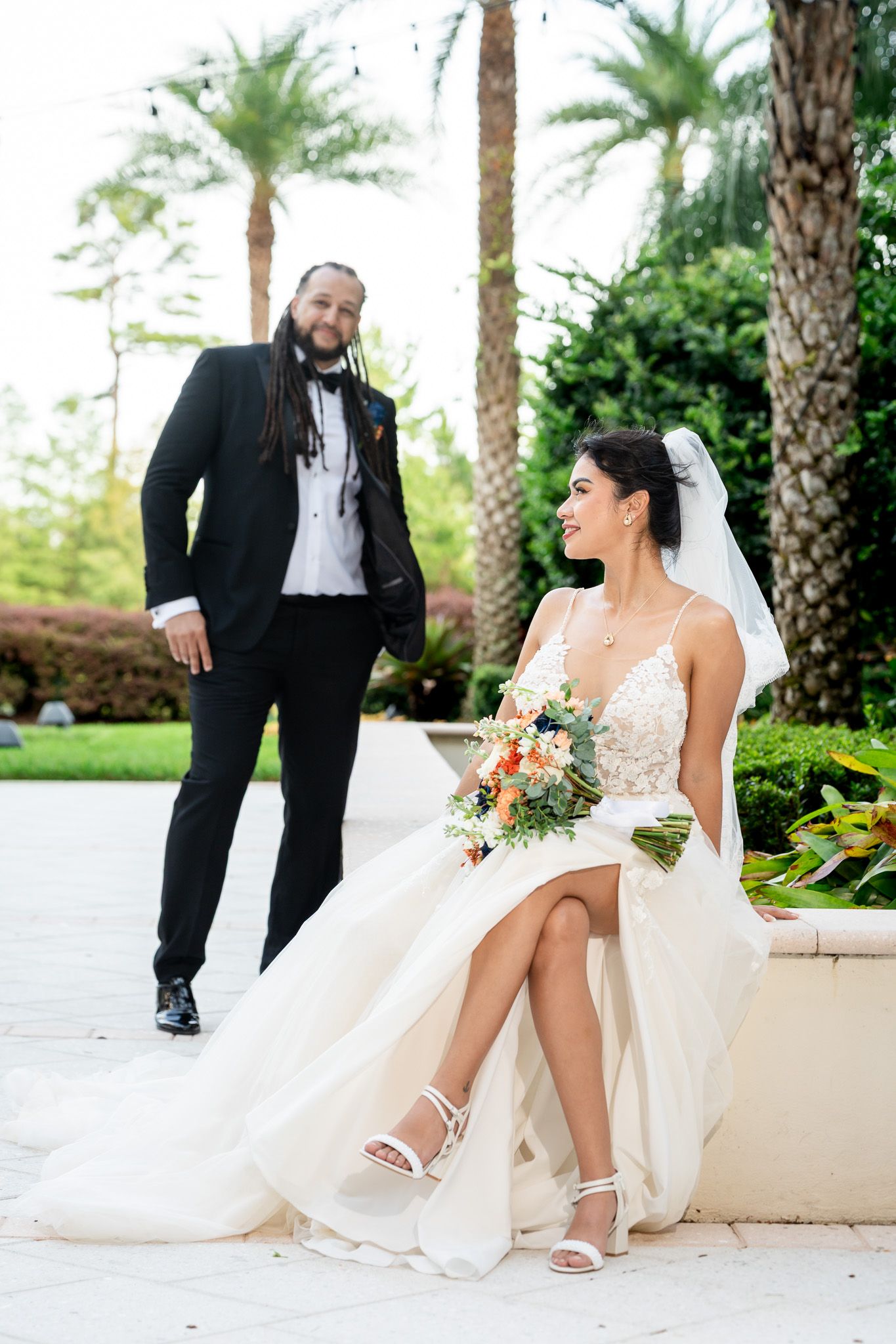 Bride seated with colorful bouquet smiling at groom approaching on palm-lined path.
