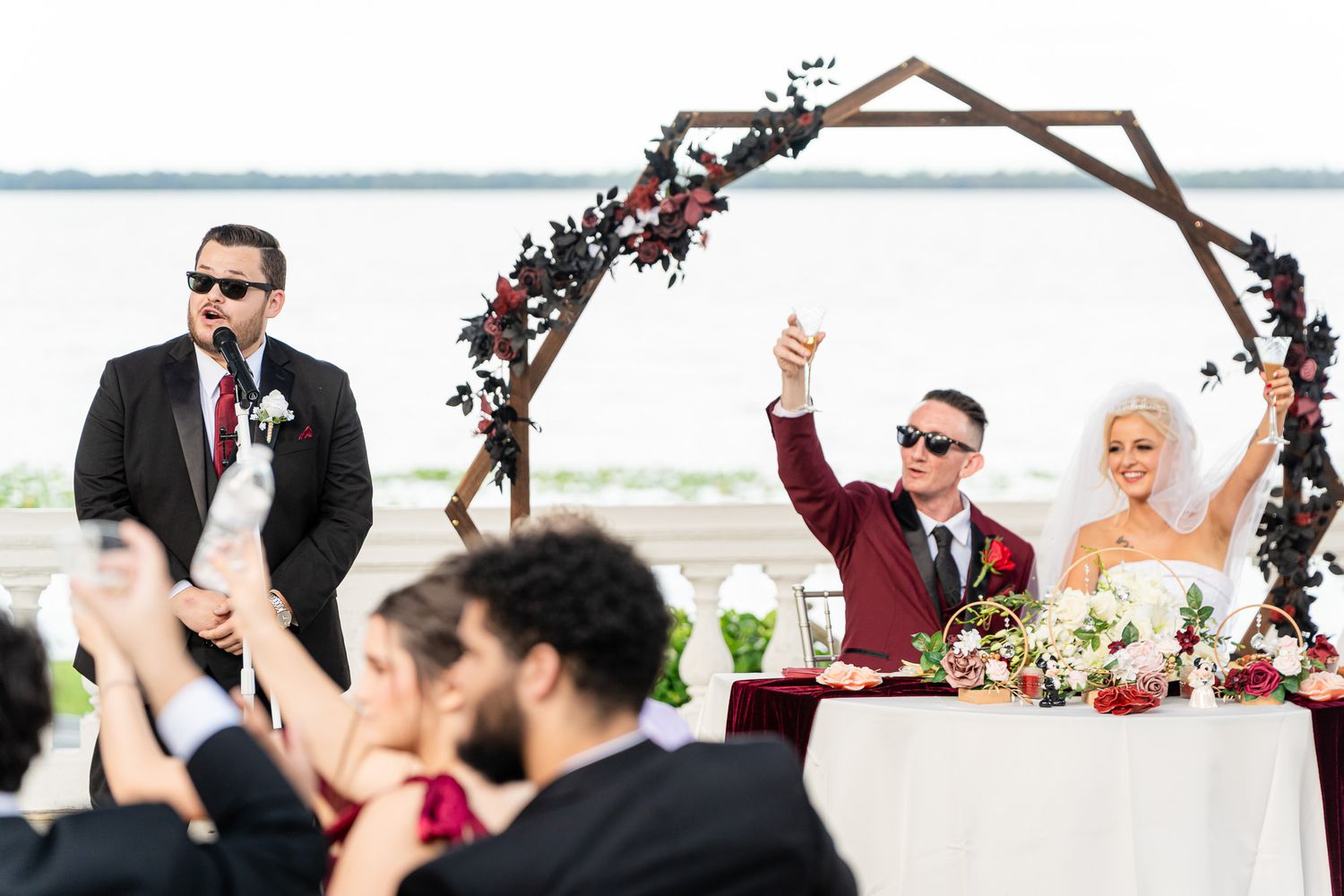 Groom in burgundy suit raising toast at outdoor waterfront wedding reception under geometric arch.