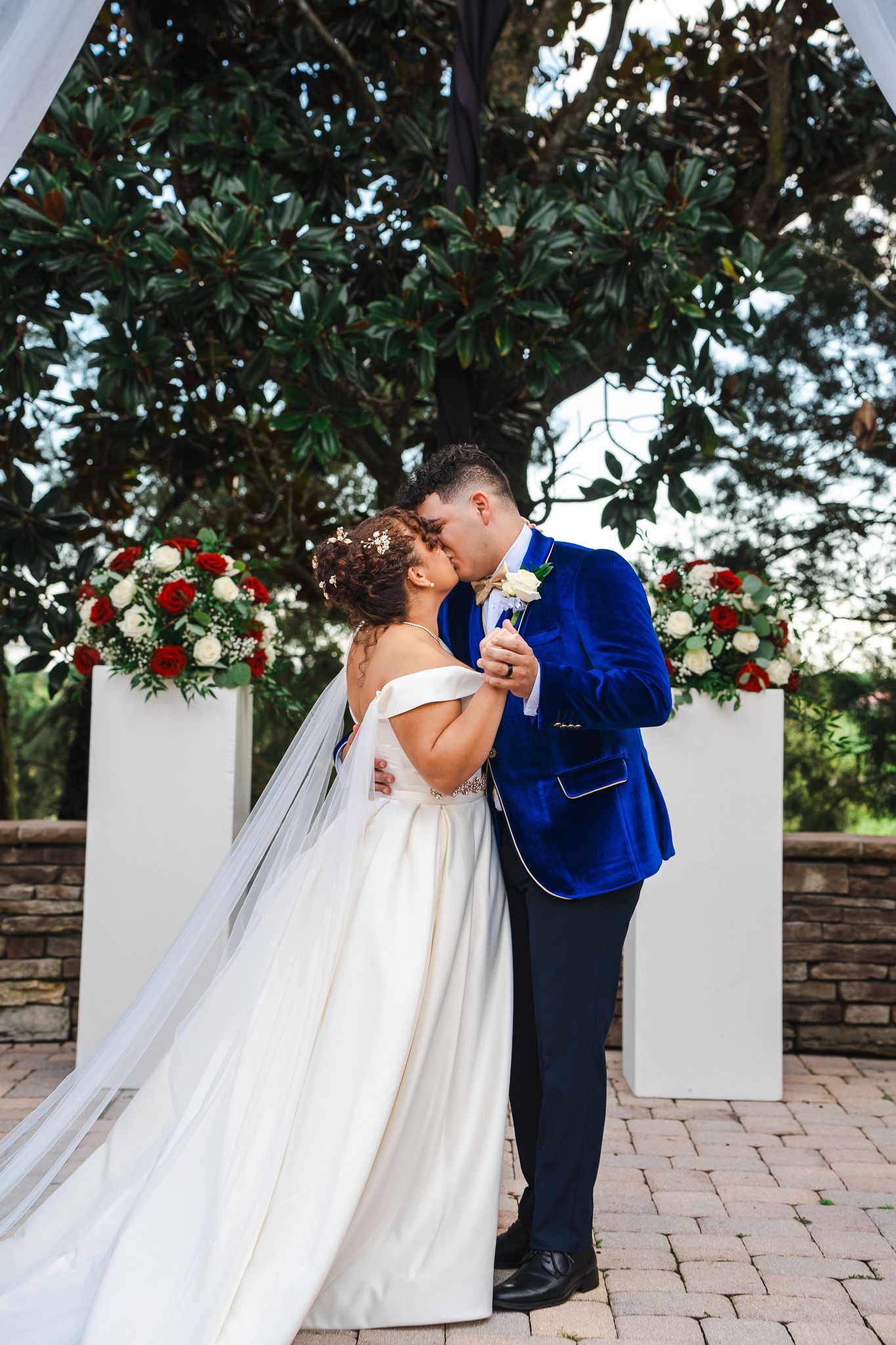 Bride and groom share a first kiss at outdoor wedding altar decorated with red and white floral arrangements.