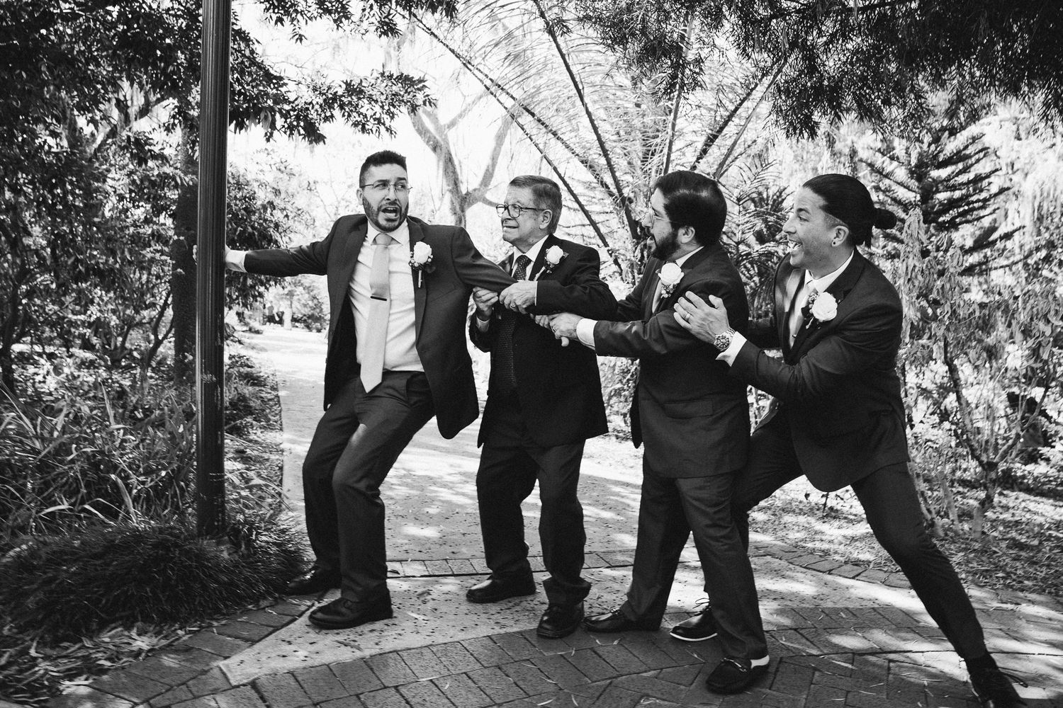 Black and white photo of groomsmen playfully posing together on a brick pathway outdoors.