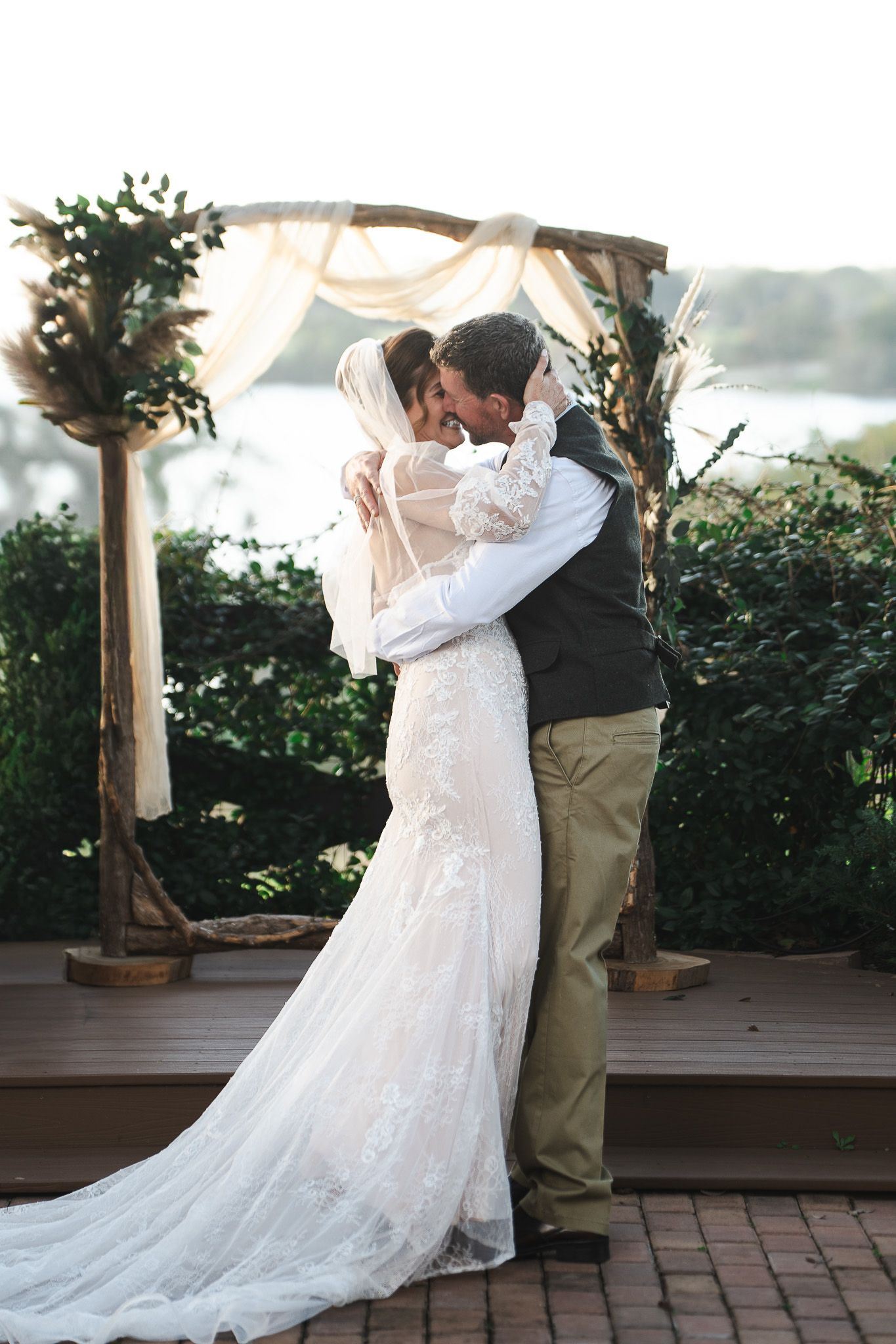 Bride and groom share first kiss under rustic wooden arch at outdoor waterfront wedding.