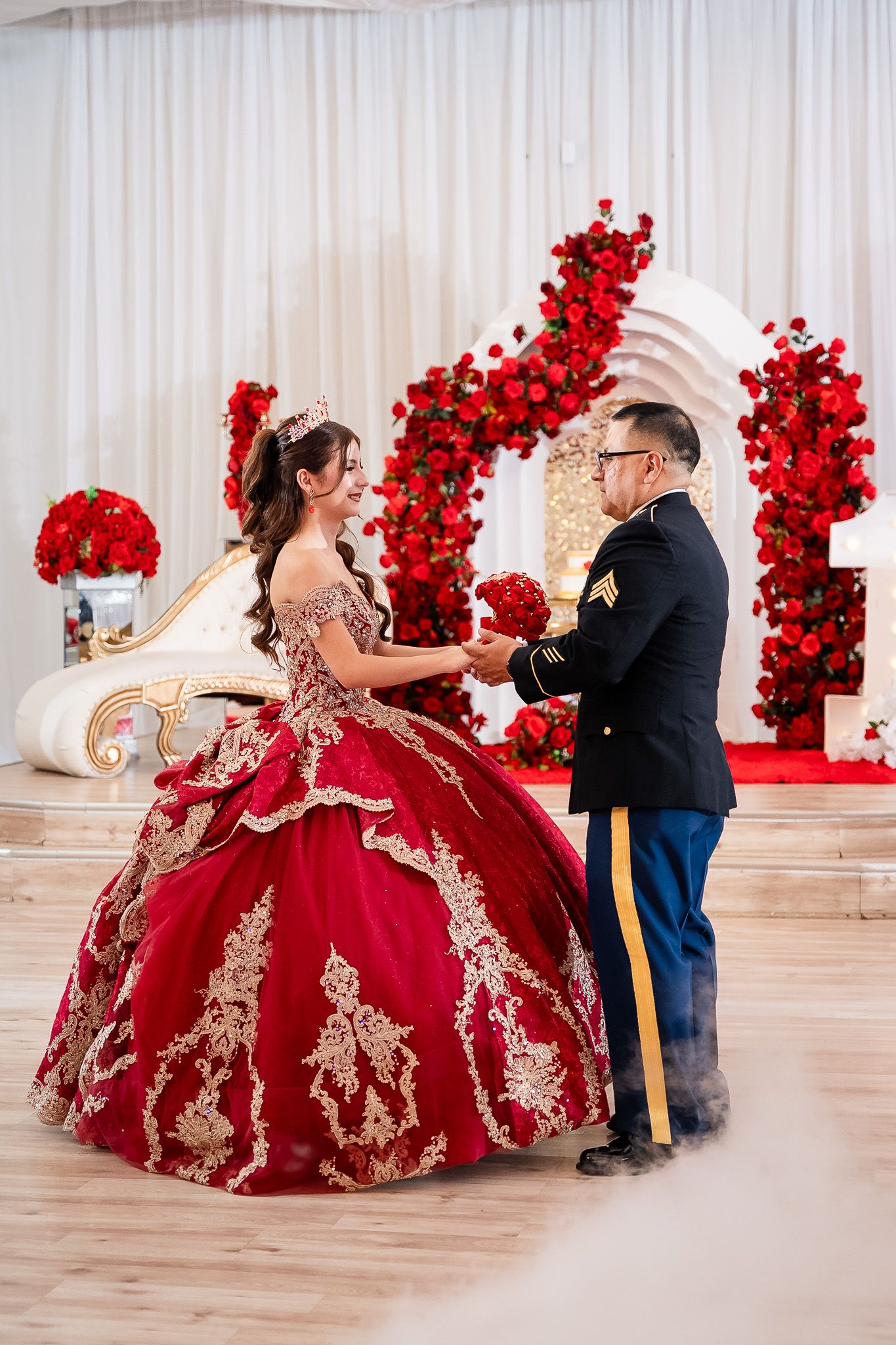 Young woman in red and gold quinceañera gown dances with man in military uniform at elegant red rose decorated venue.