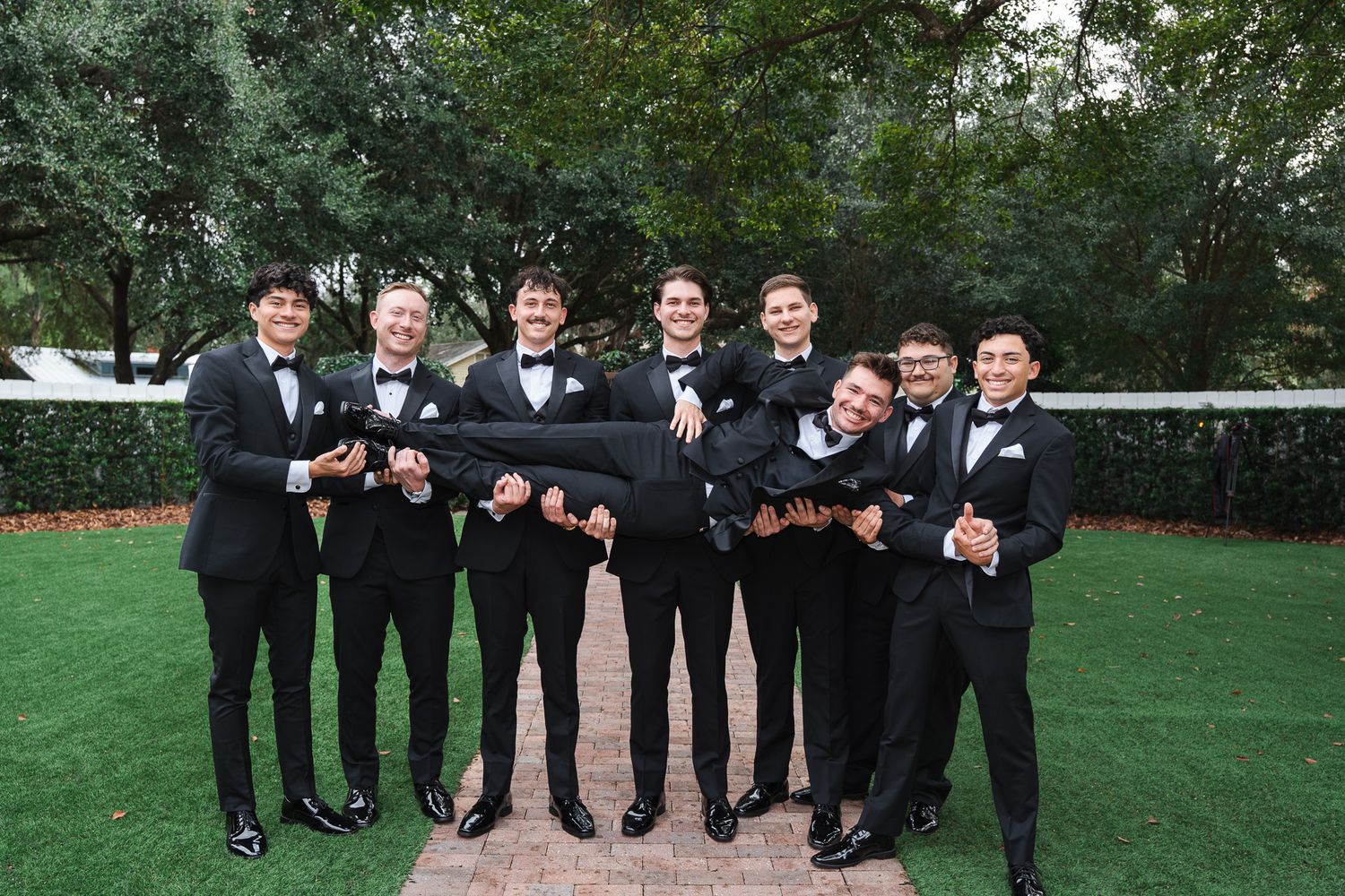 Groomsmen in black tuxedos playfully lift the groom horizontally outdoors before the wedding.