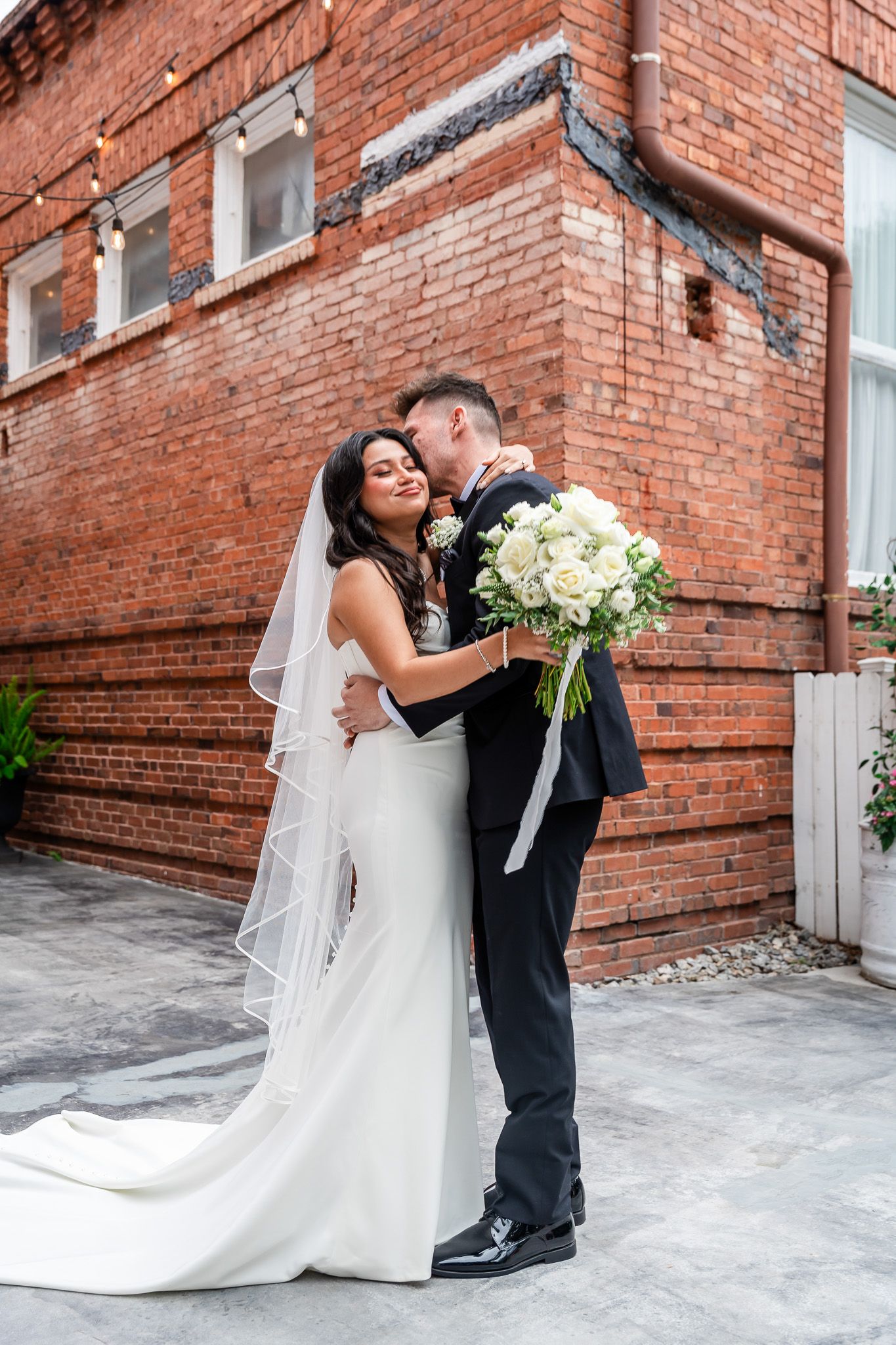 Bride and groom share a romantic embrace in front of a rustic red brick wall with white rose bouquet.