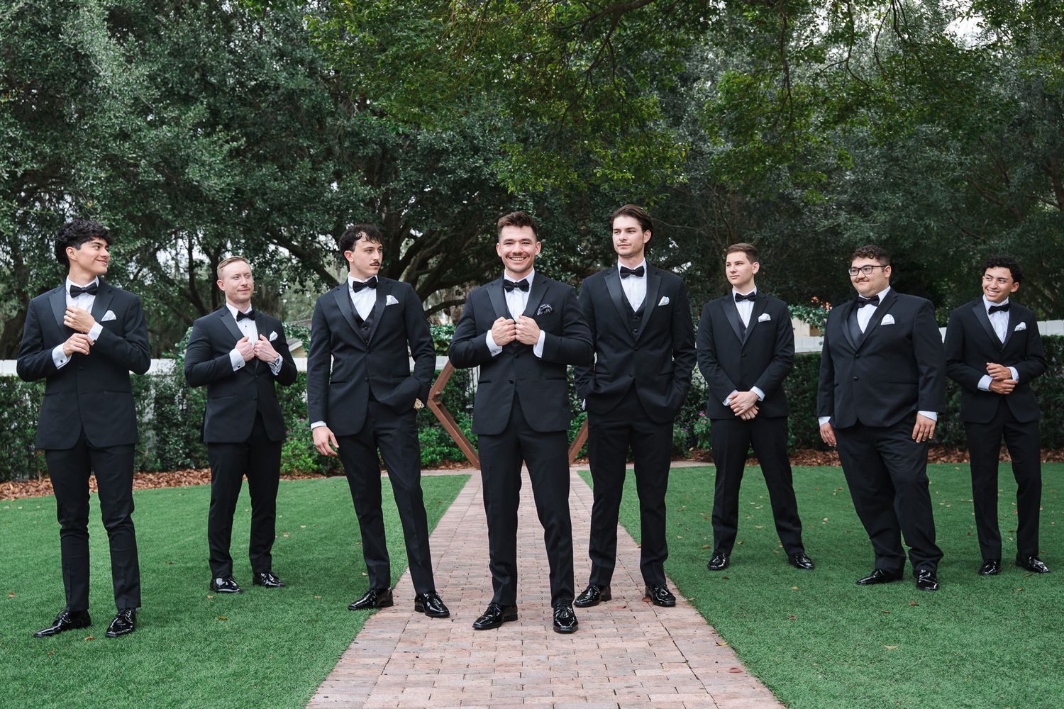 Groom and groomsmen in black tuxedos posing on brick pathway surrounded by greenery.