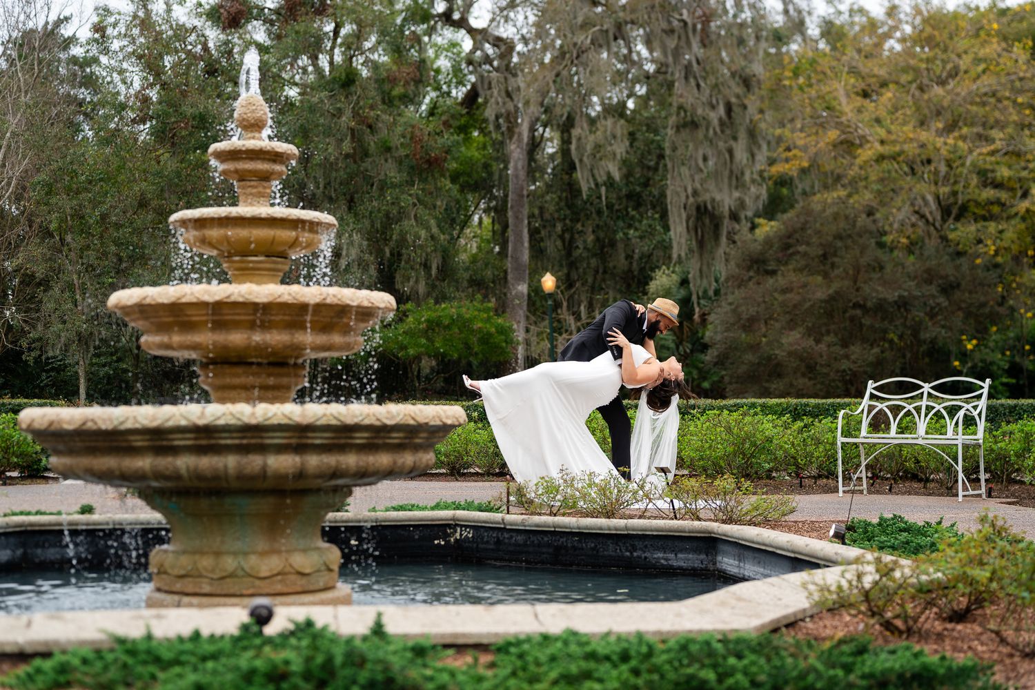 Bride and groom share a romantic dip kiss beside an elegant stone fountain in a garden setting.