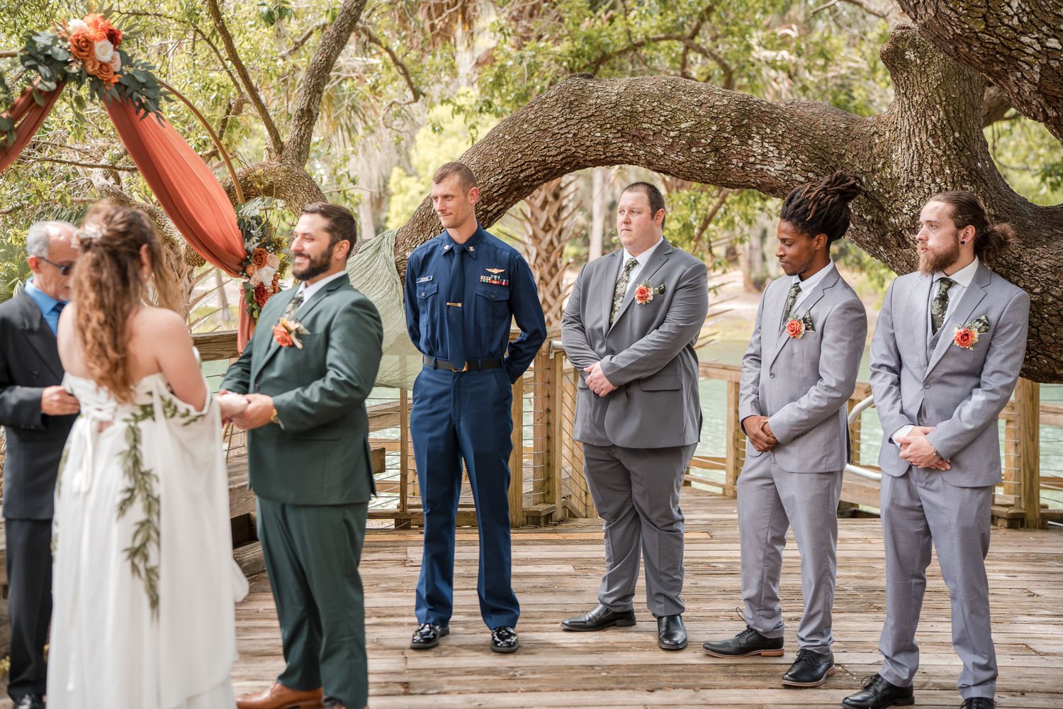 Outdoor wedding ceremony on wooden deck with bride, groom, officiant in Coast Guard uniform, and groomsmen under oak tree.