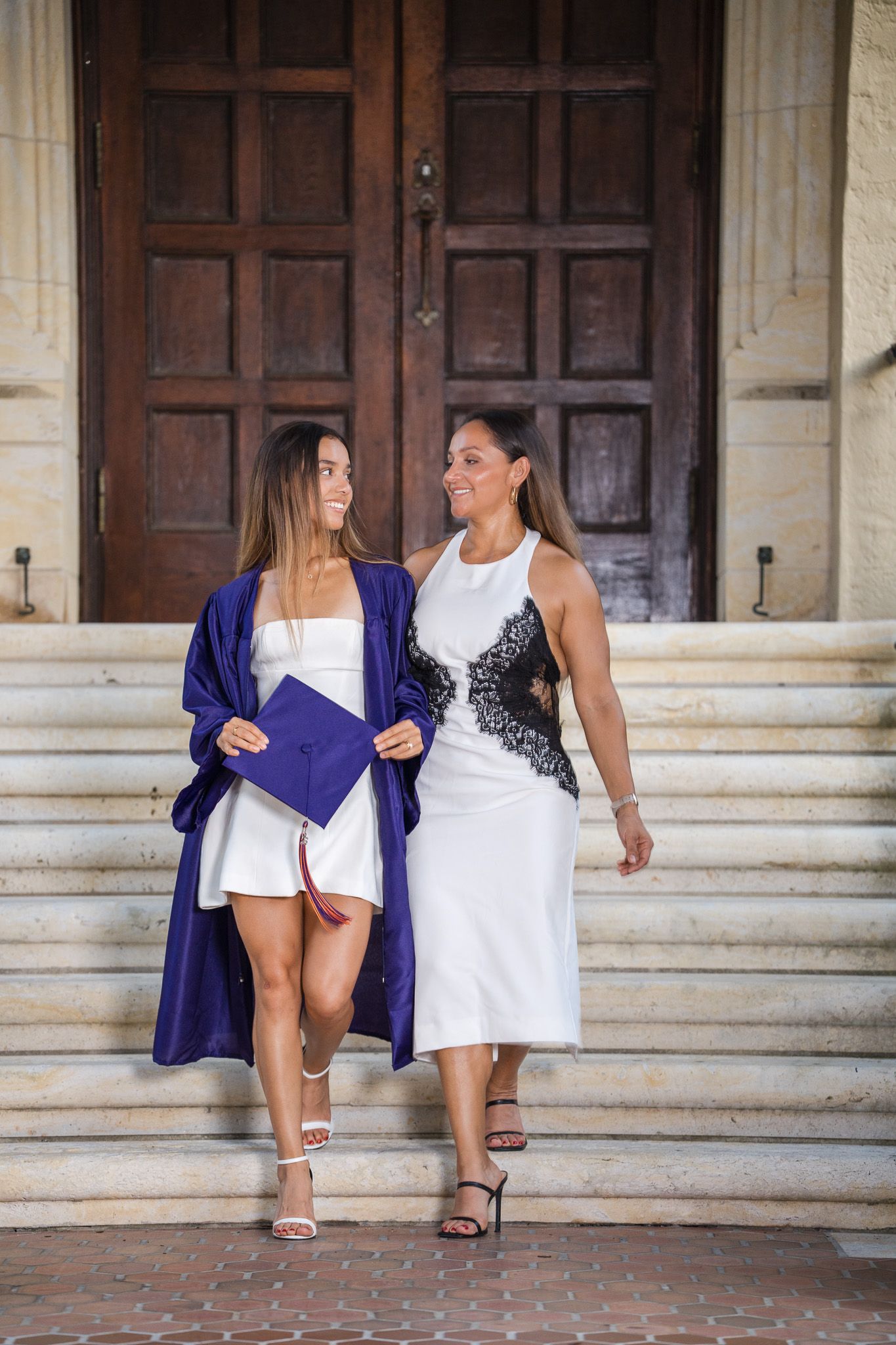 Graduate in blue cap and gown walking down steps with woman in white dress at building entrance.