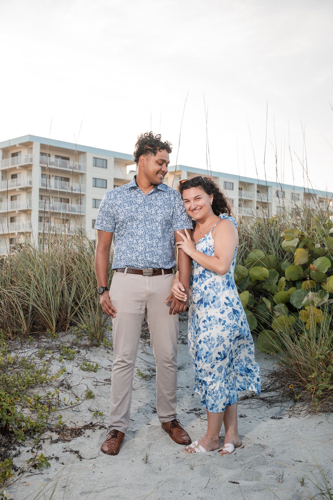 Couple in matching blue floral outfits smiling together on sandy beach dunes with oceanfront hotel in background.
