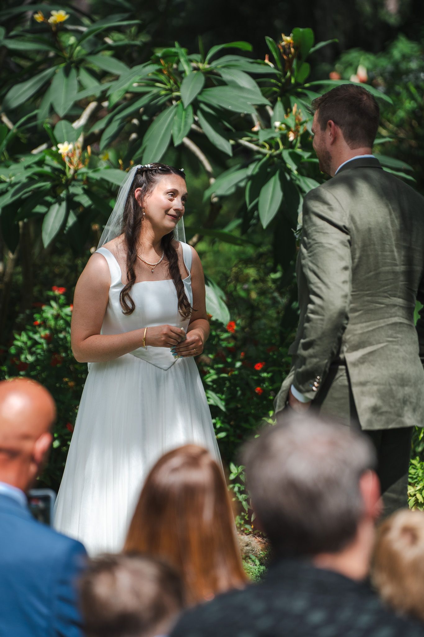 Radiant bride smiles while reciting vows to groom at lush tropical outdoor wedding ceremony.
