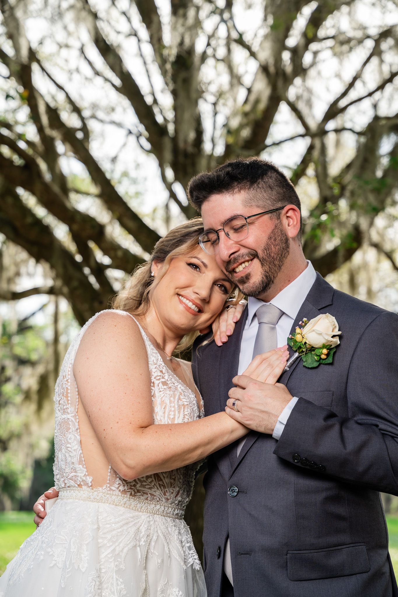 Smiling bride in lace gown leans against groom in gray suit with boutonniere under a large moss-draped oak tree.