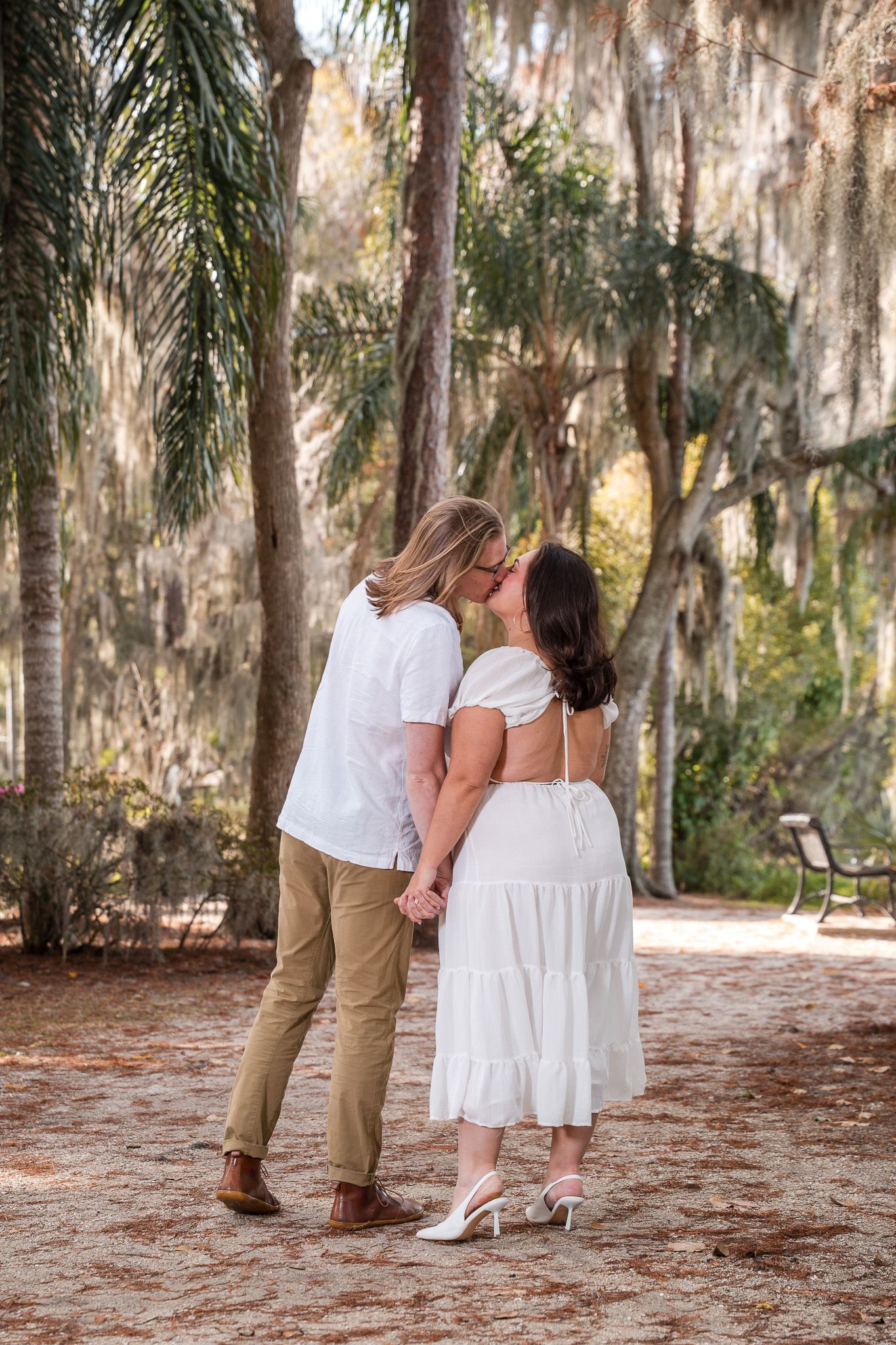 Couple sharing a kiss while holding hands on a sandy path among tall trees.