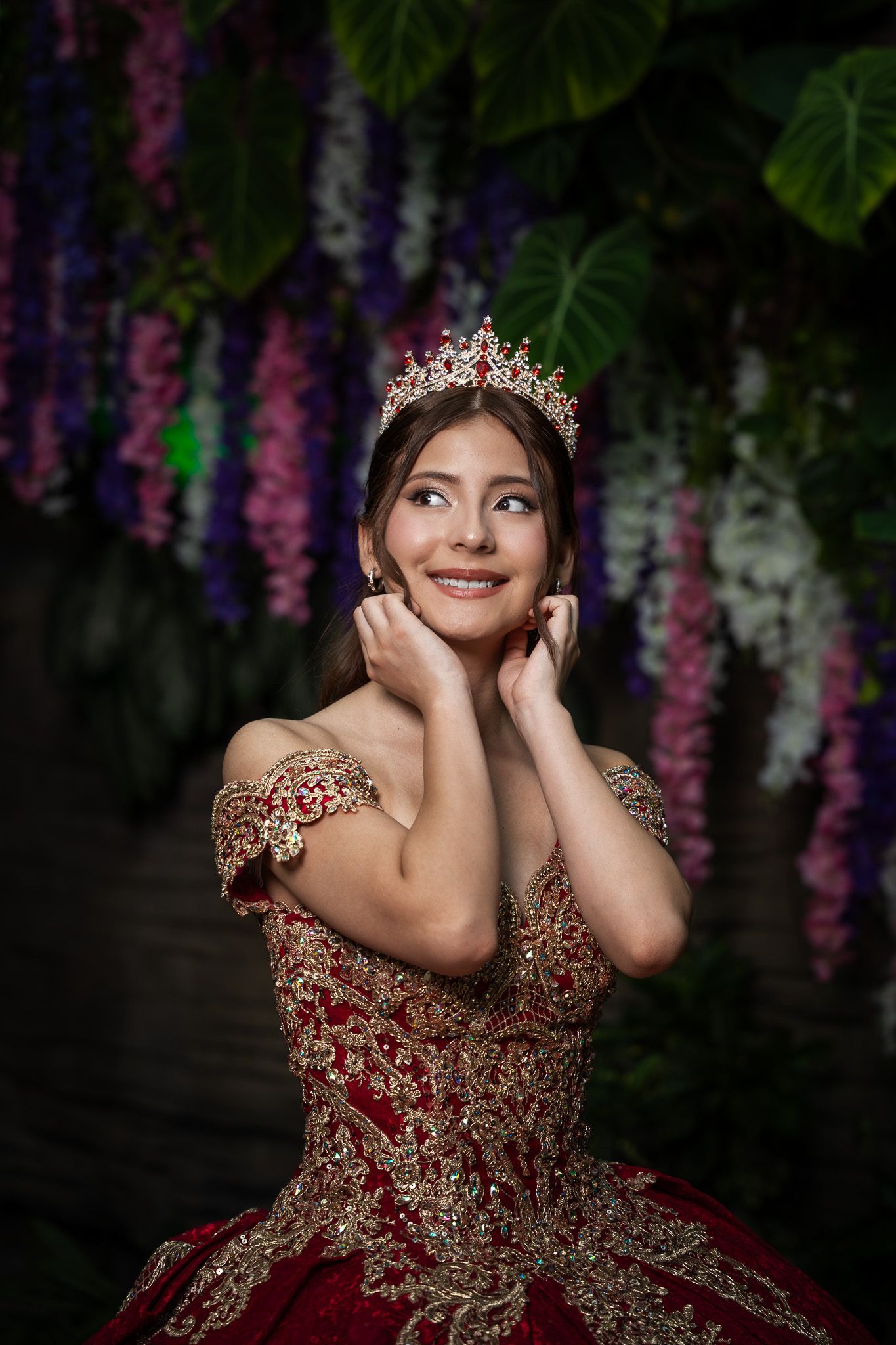Close-up of smiling young woman wearing red gold quinceañera dress and jeweled crown surrounded by hanging wisteria blooms.