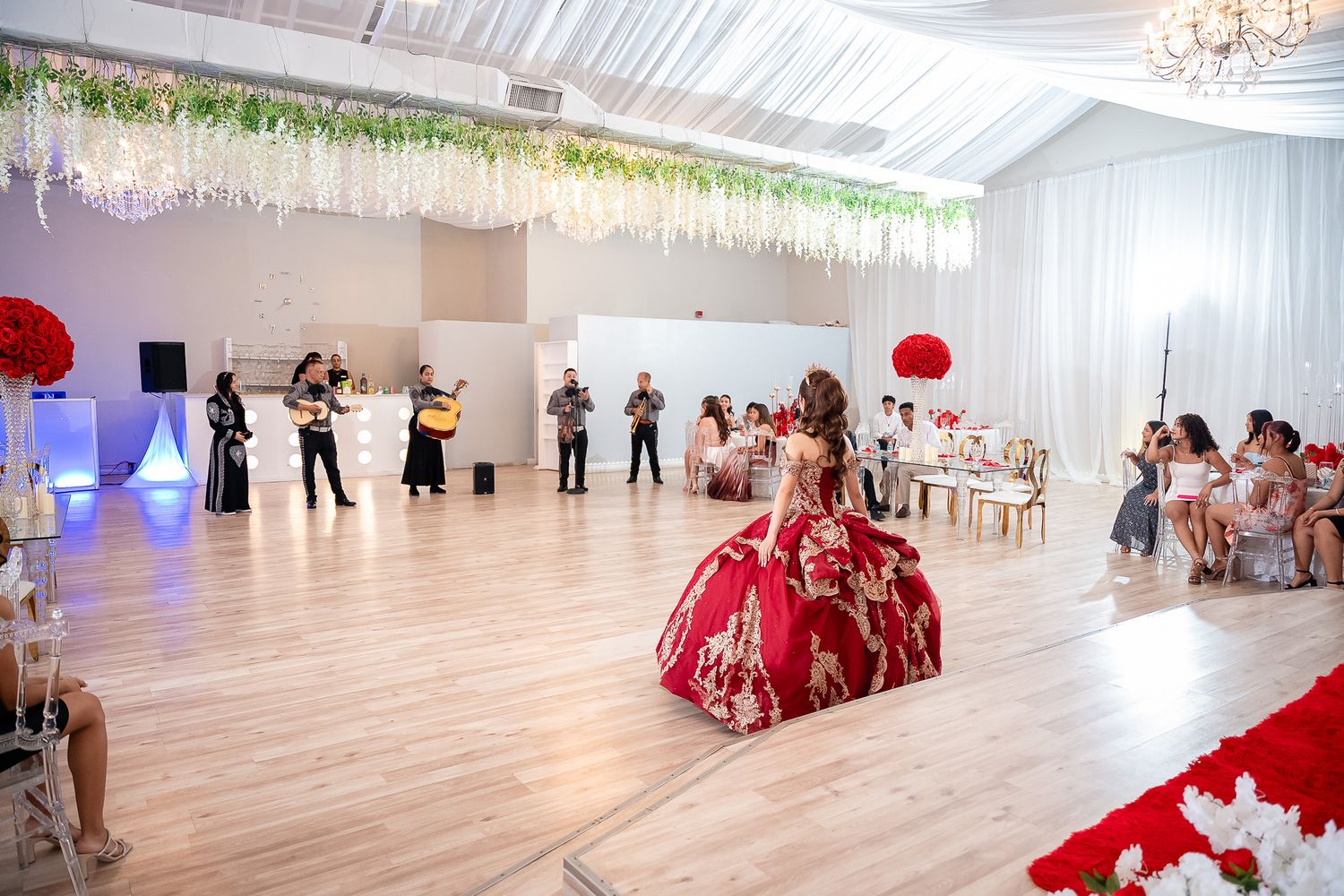A girl in a red quinceañera gown dances at an elegant venue with live musicians and floral decorations.