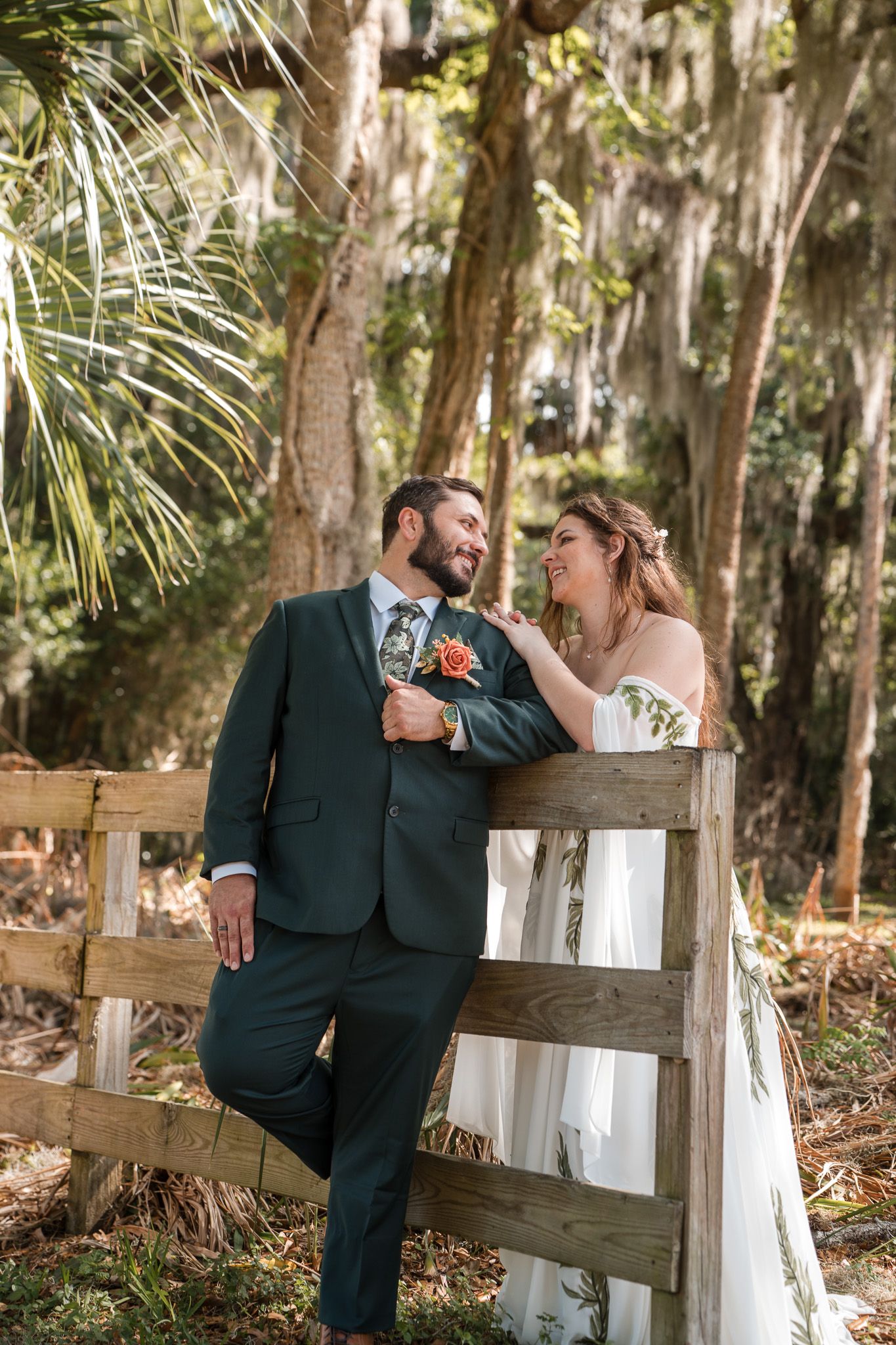 Bride and groom lean against rustic wooden fence surrounded by lush trees with Spanish moss at outdoor wedding.