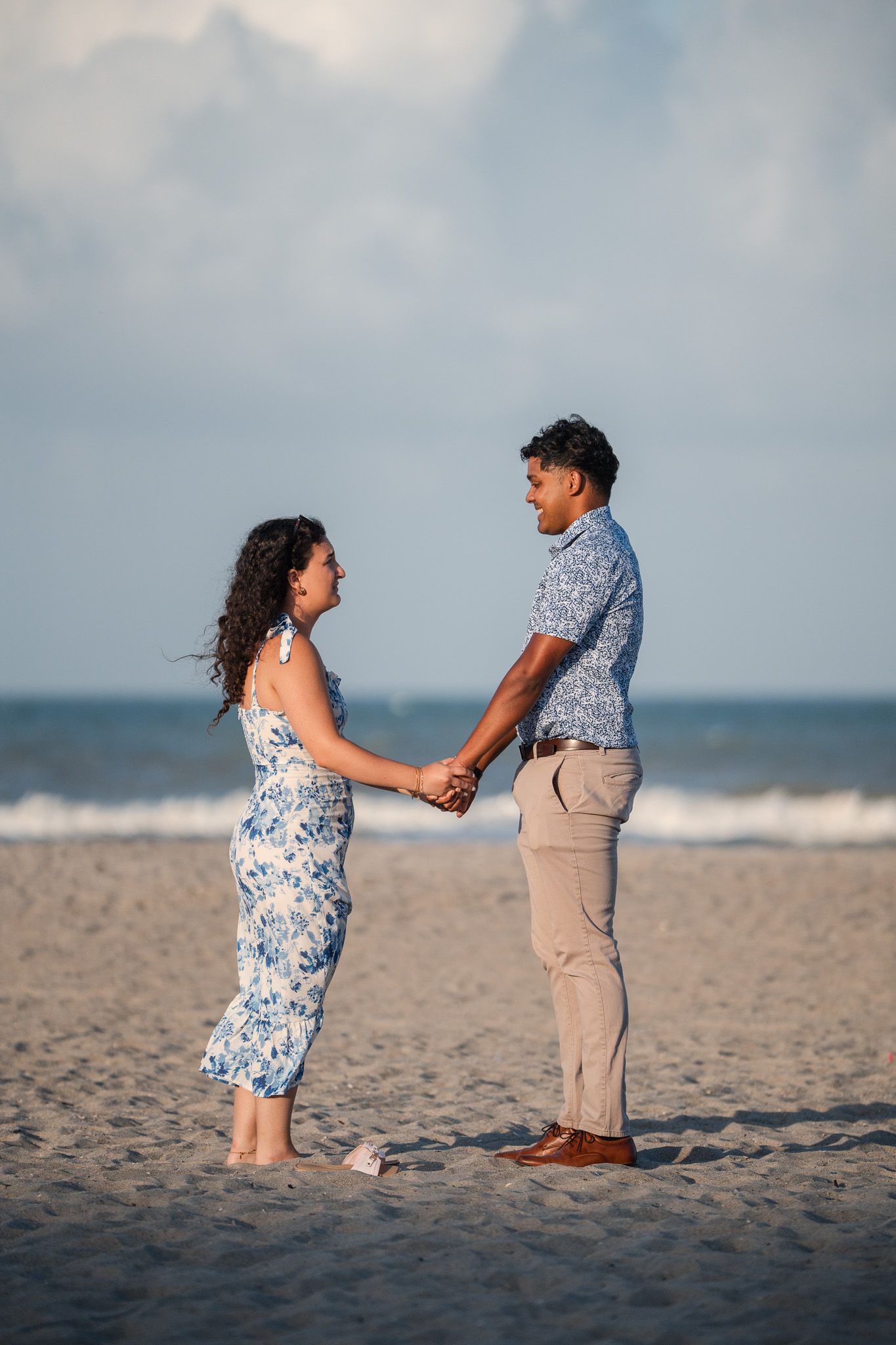Couple holding hands and smiling at each other on a sandy beach with ocean waves in the background.