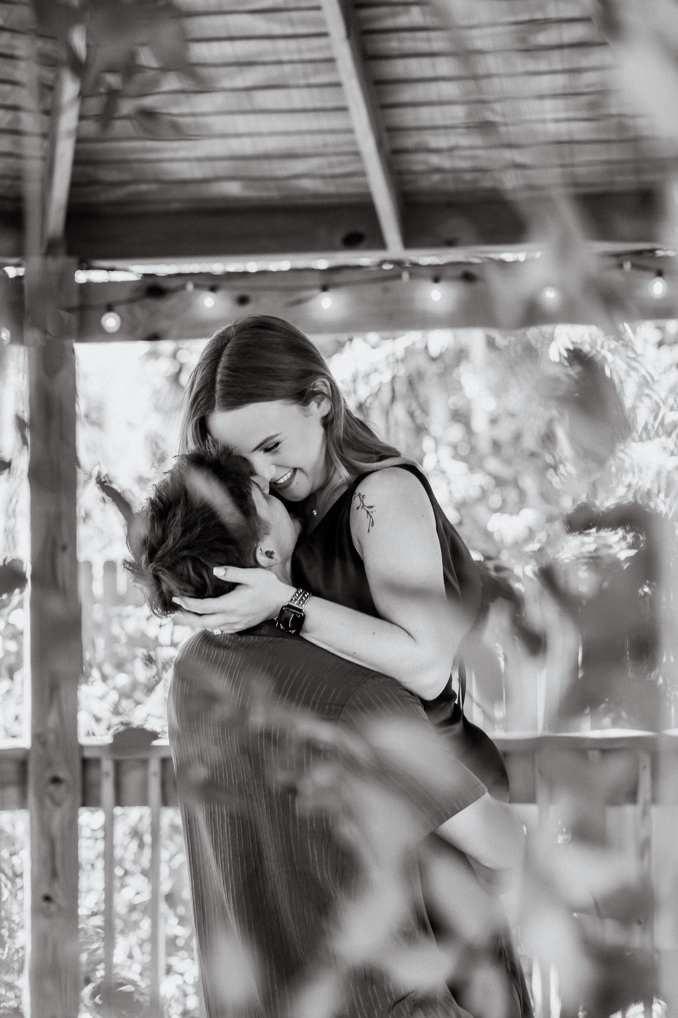 Black and white photo of a couple embracing tenderly in a rustic gazebo with string lights and lush greenery.