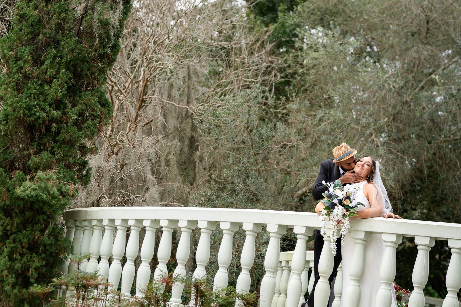 Bride and groom share tender kiss leaning over ornate white balustrade railing in garden setting.
