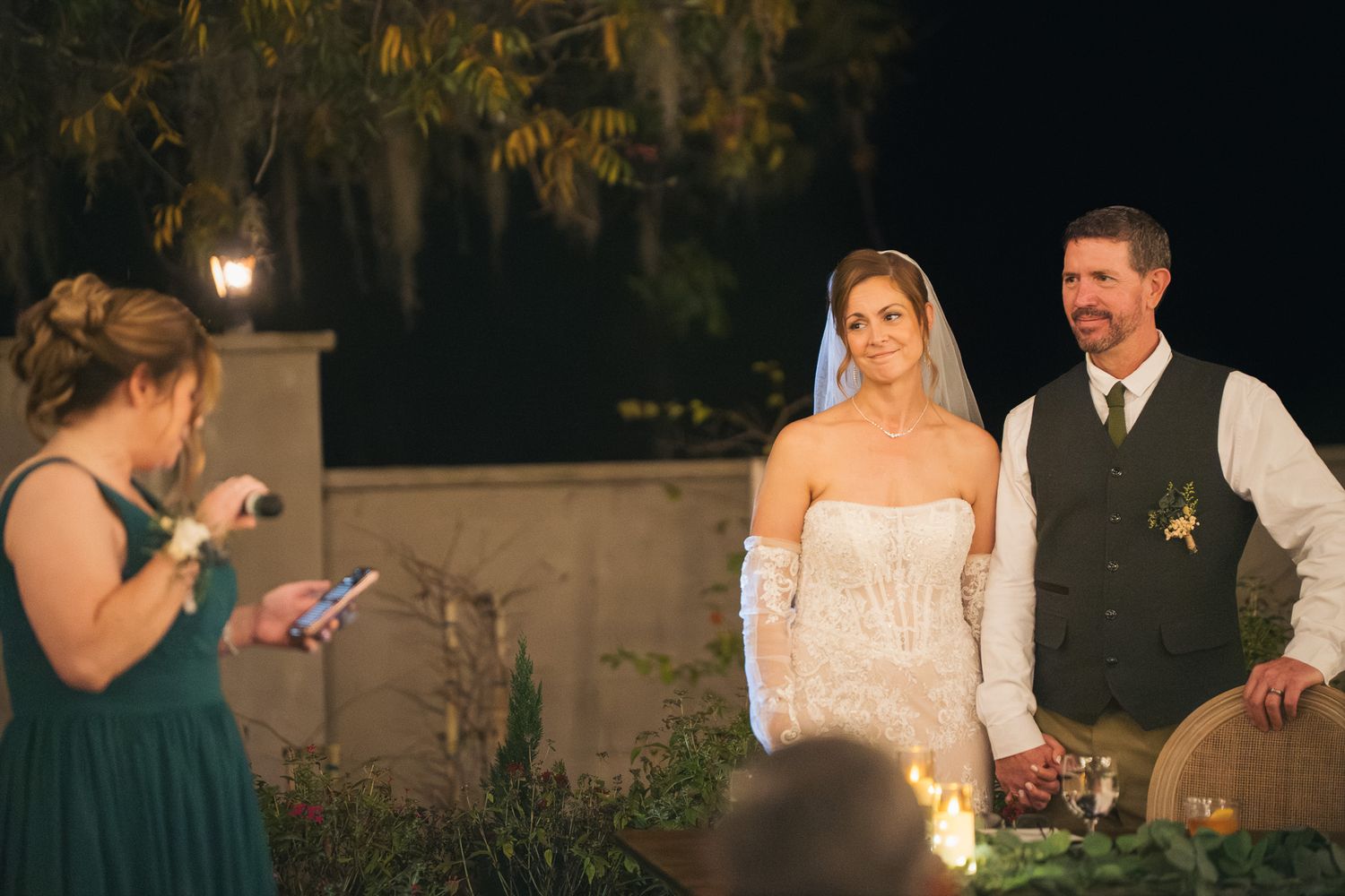 Bride and groom hold hands during their outdoor evening wedding ceremony with a female officiant.