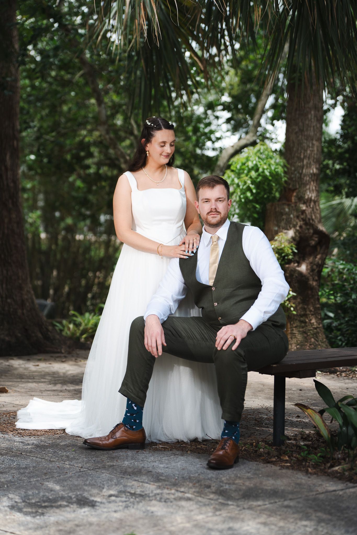 Wedding couple portrait with groom seated on bench and bride standing in garden.
