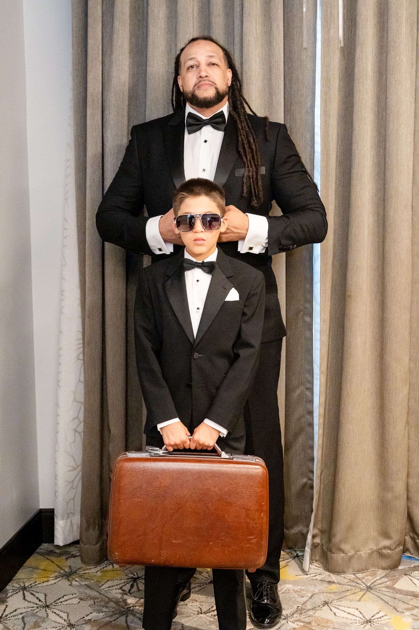 Groom in tuxedo places sunglasses on young boy in matching suit holding a vintage briefcase.