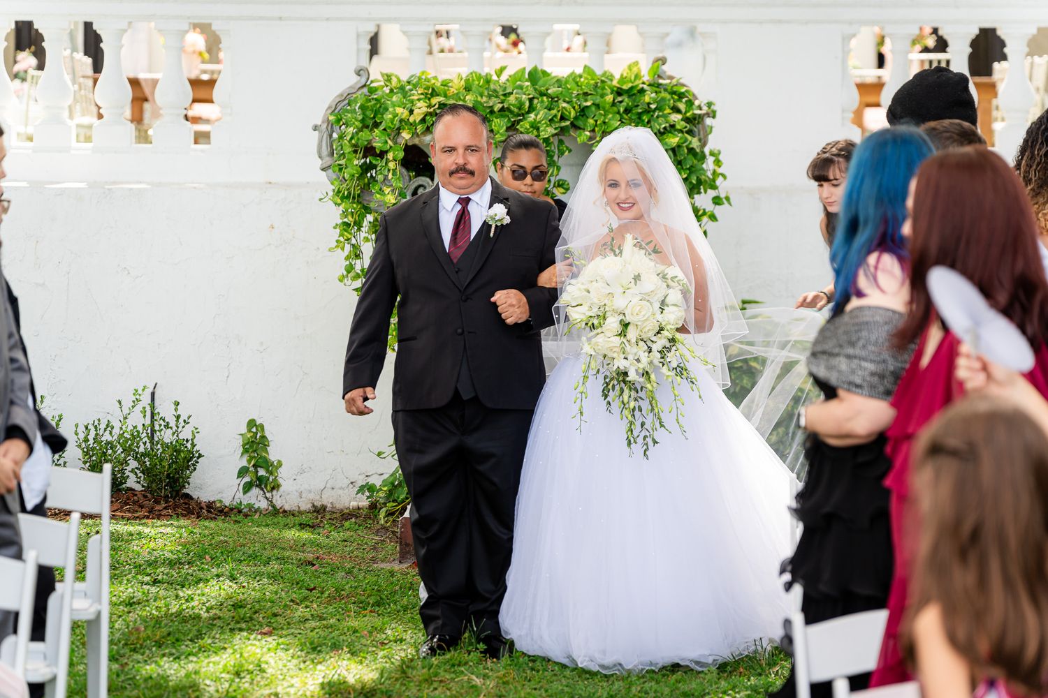 Father escorts radiant bride in white ball gown down outdoor wedding ceremony aisle.