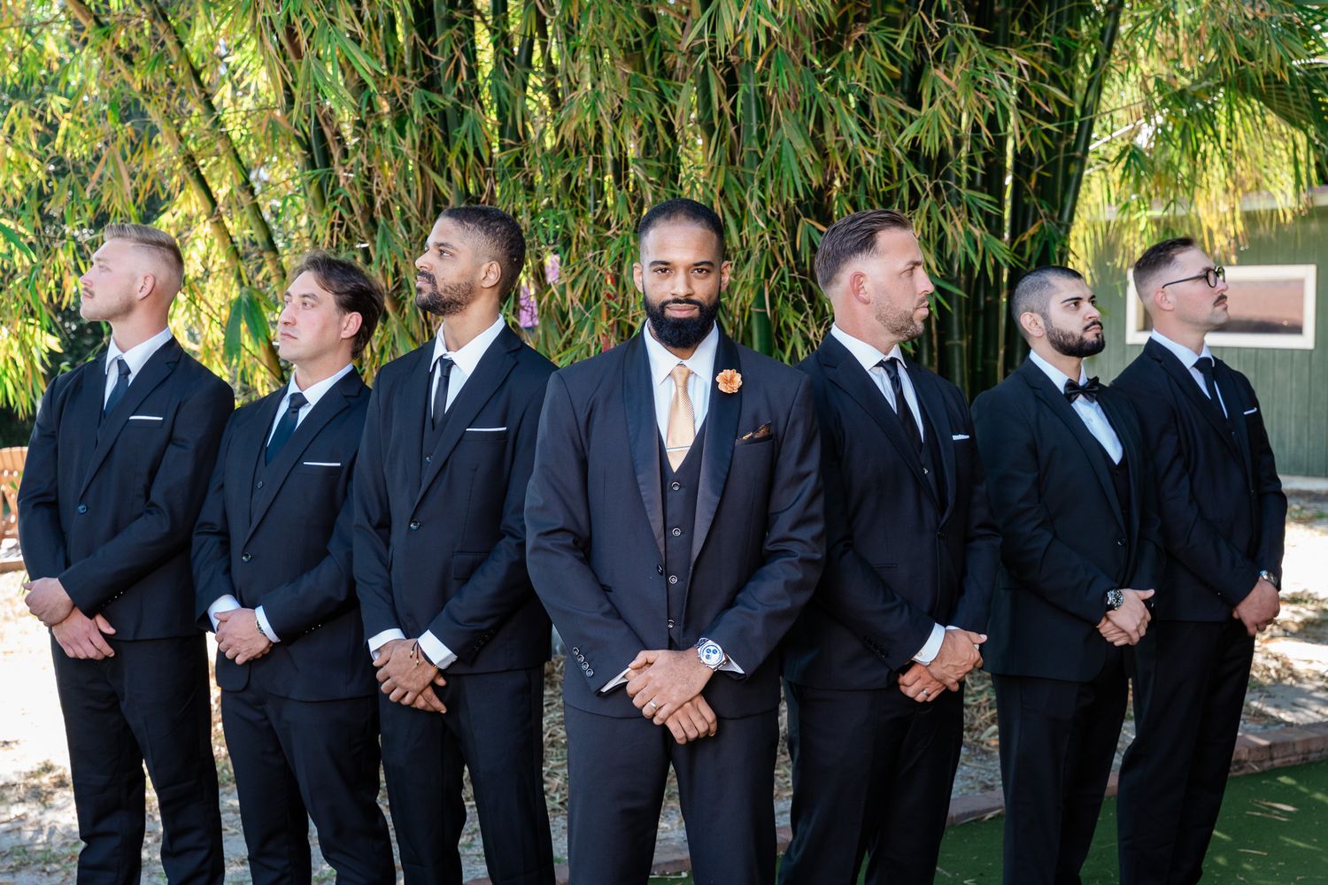 Groom in navy suit stands centered with groomsmen in black tuxedos lined up behind him outdoors.
